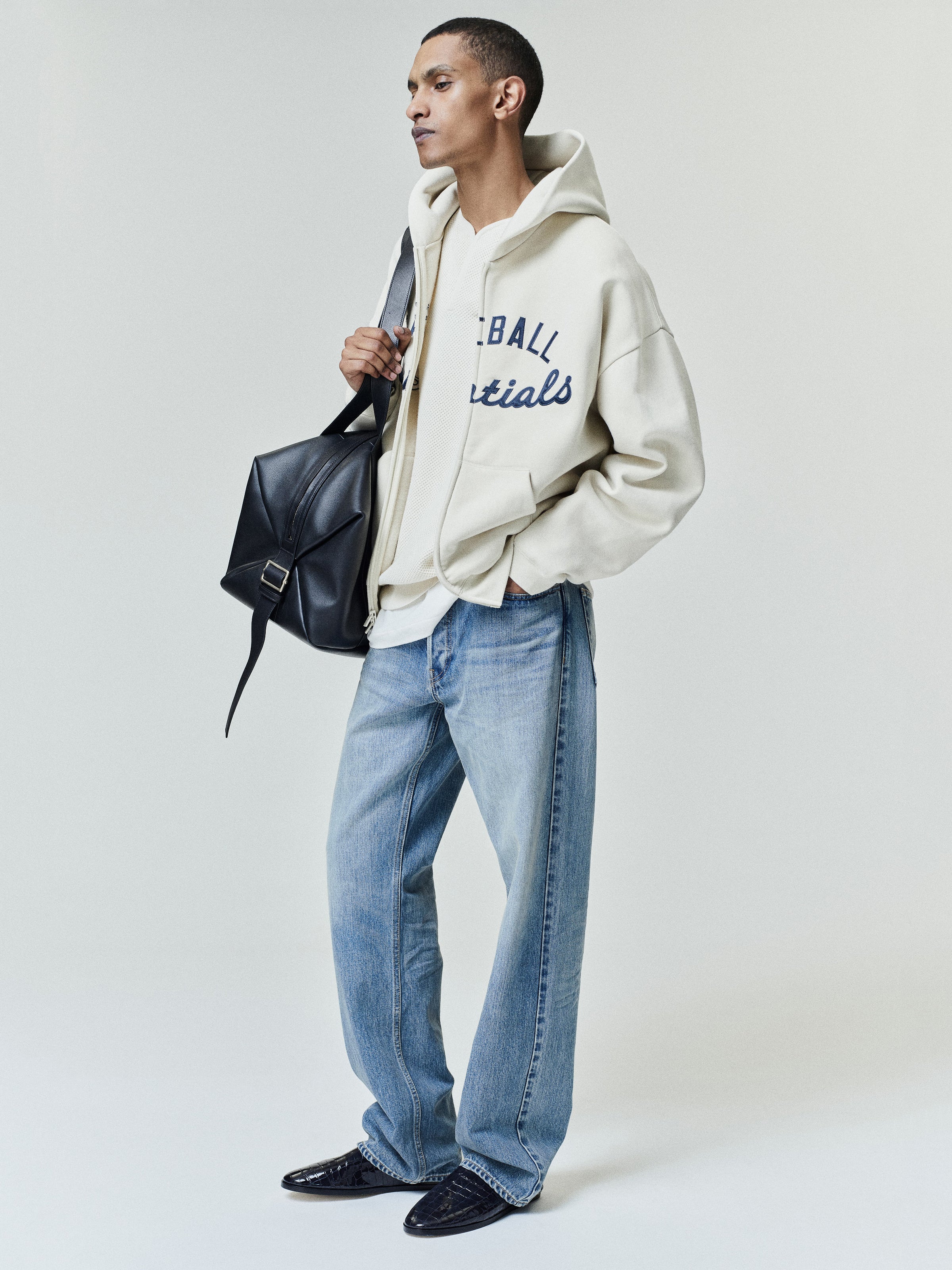 Model wearing an off-white Mlb Full Zip Sport Hoodie with navy script detailing, paired with light blue jeans and black leather shoes, accessorized with a black leather shoulder bag.