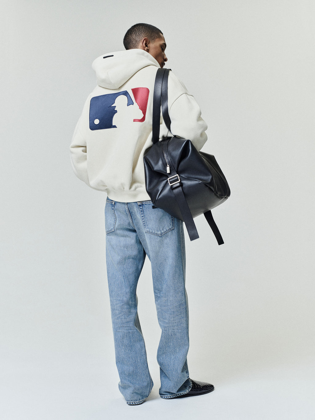 Model wearing an off-white Mlb Full Zip Sport Hoodie with a large red and blue graphic on the back, paired with light blue jeans and carrying a black leather backpack.