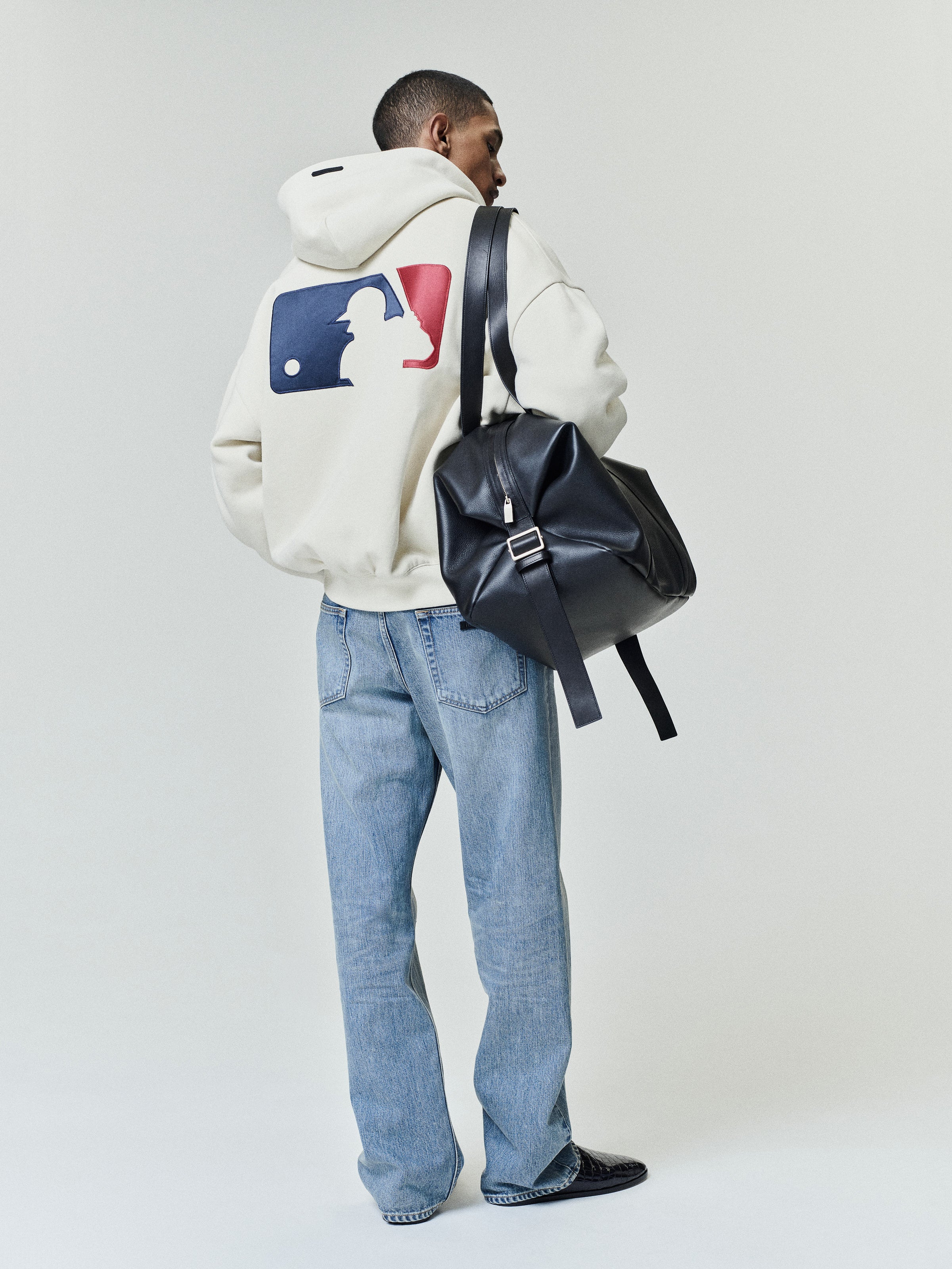 Model wearing an off-white Mlb Full Zip Sport Hoodie with a large red and blue graphic on the back, paired with light blue jeans and carrying a black leather backpack.