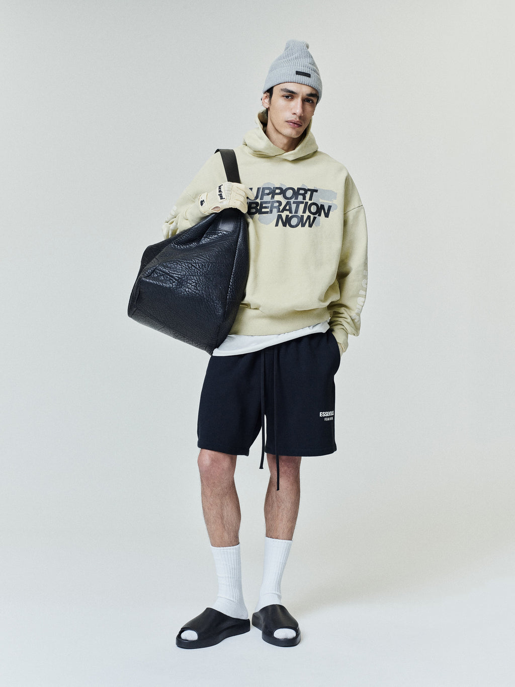 Man wearing a light beige hoodie with graphic text, black shorts, white socks, black slide sandals, gray beanie, and holding a large black leather bag against a plain background