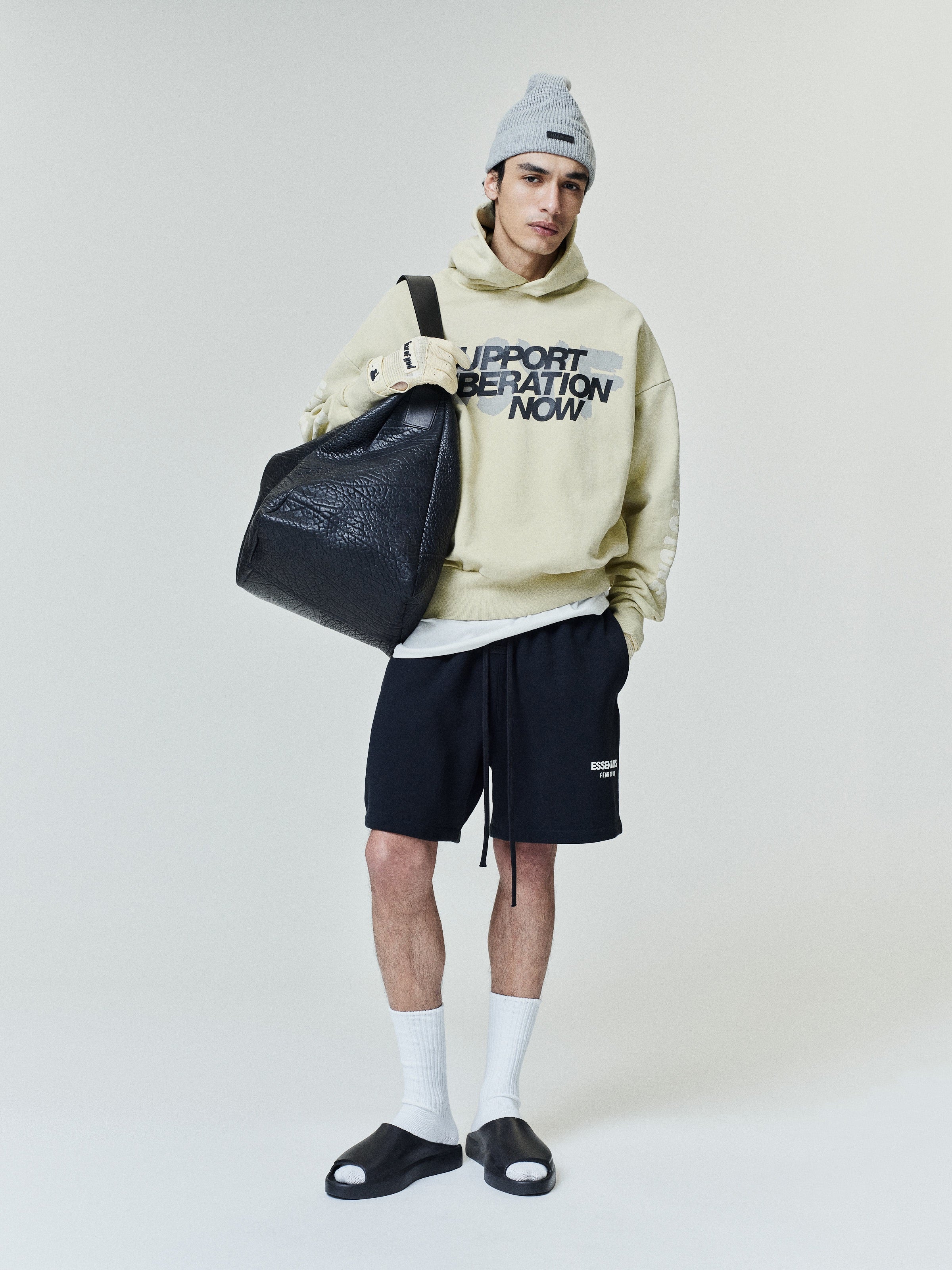 Man wearing a light beige hoodie with graphic text, black shorts, white socks, black slide sandals, gray beanie, and holding a large black leather bag against a plain background