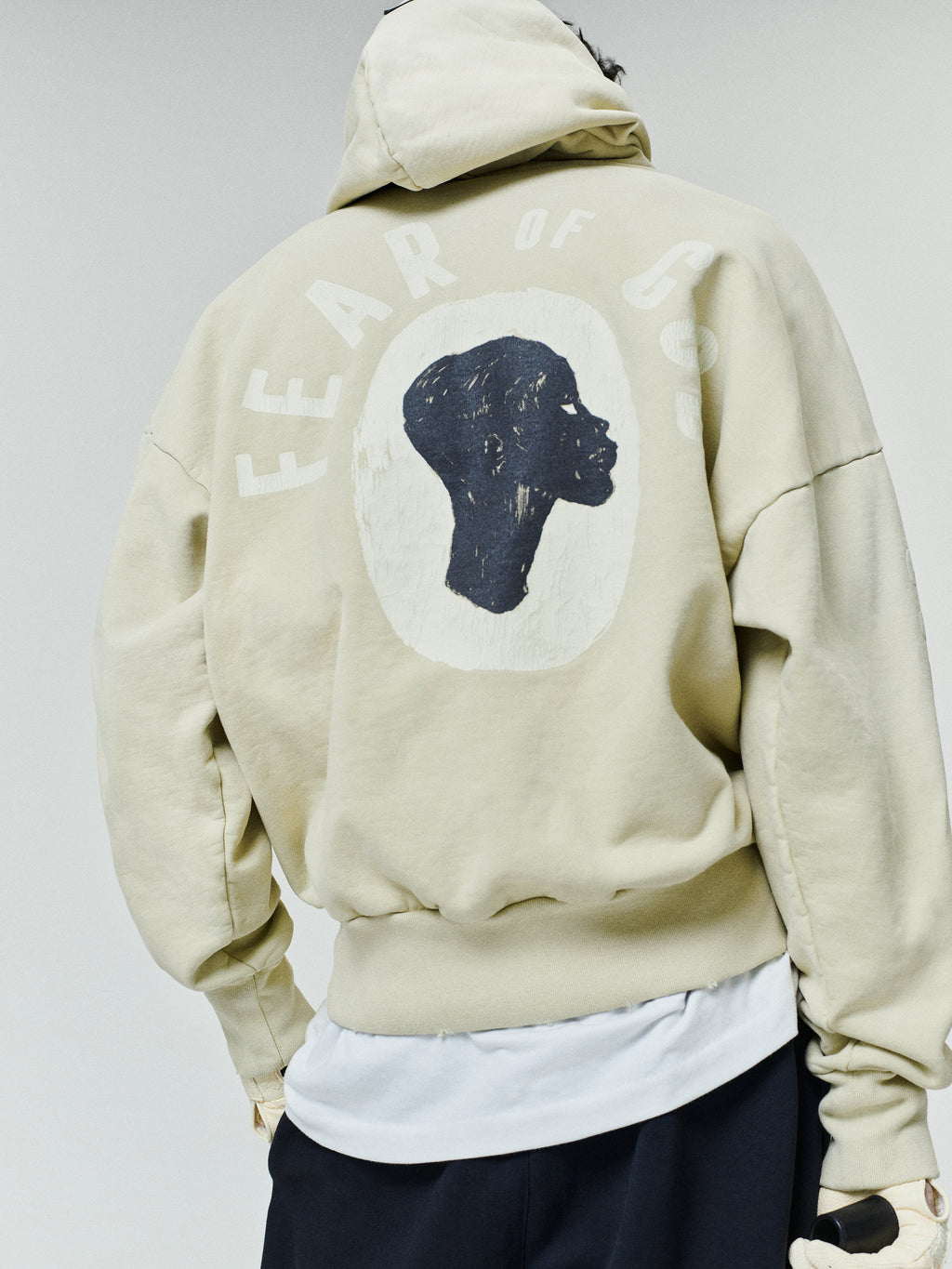 Person wearing an oversized light beige hoodie with a black silhouette profile graphic and "Fear of God" text printed on the back, paired with black pants and a white shirt underneath