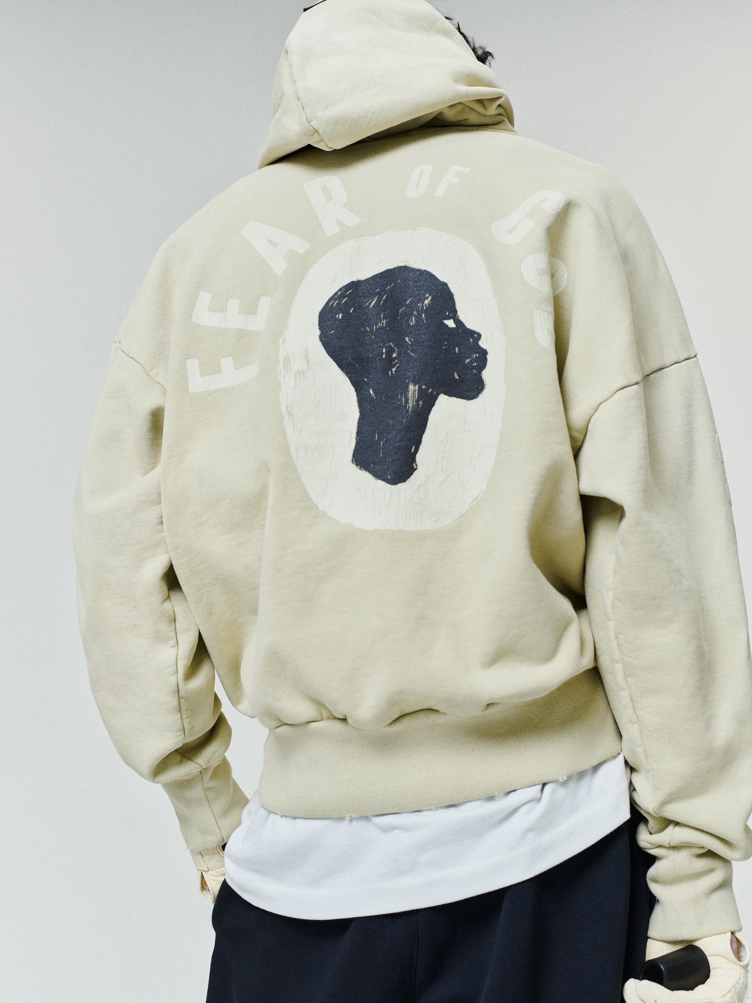 Person wearing an oversized light beige hoodie with a black silhouette profile graphic and "Fear of God" text printed on the back, paired with black pants and a white shirt underneath