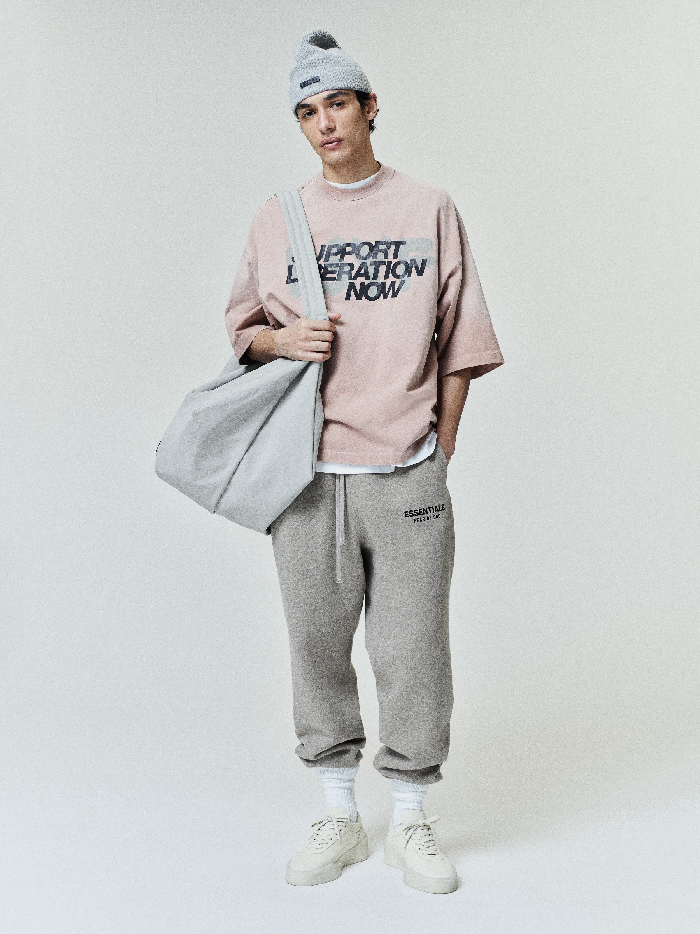 Young man wearing a loose dusty pink t-shirt with bold black text on the front, paired with light gray sweatpants, white sneakers, white socks, a light gray beanie, and carrying a large gray tote bag over his shoulder.