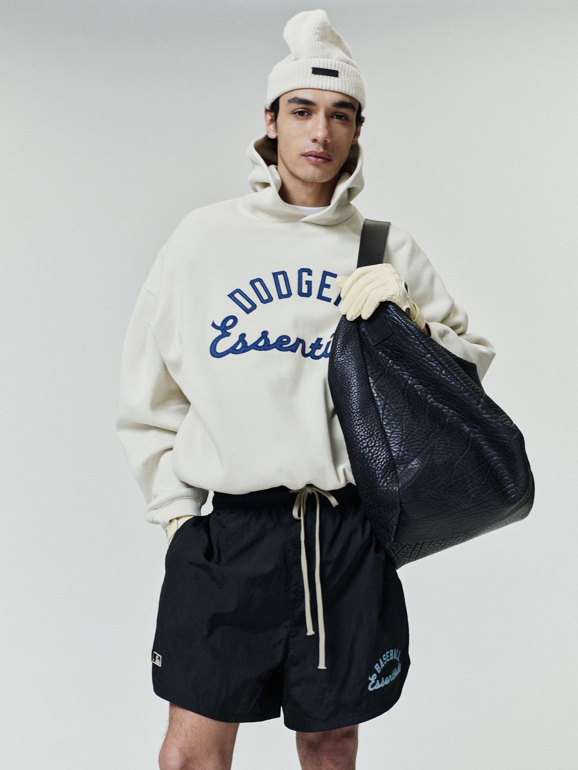 Model wearing a cream Dodgers Sport Hoodie with navy blue printed lettering, paired with black shorts and a white beanie, holding a large textured black tote bag.