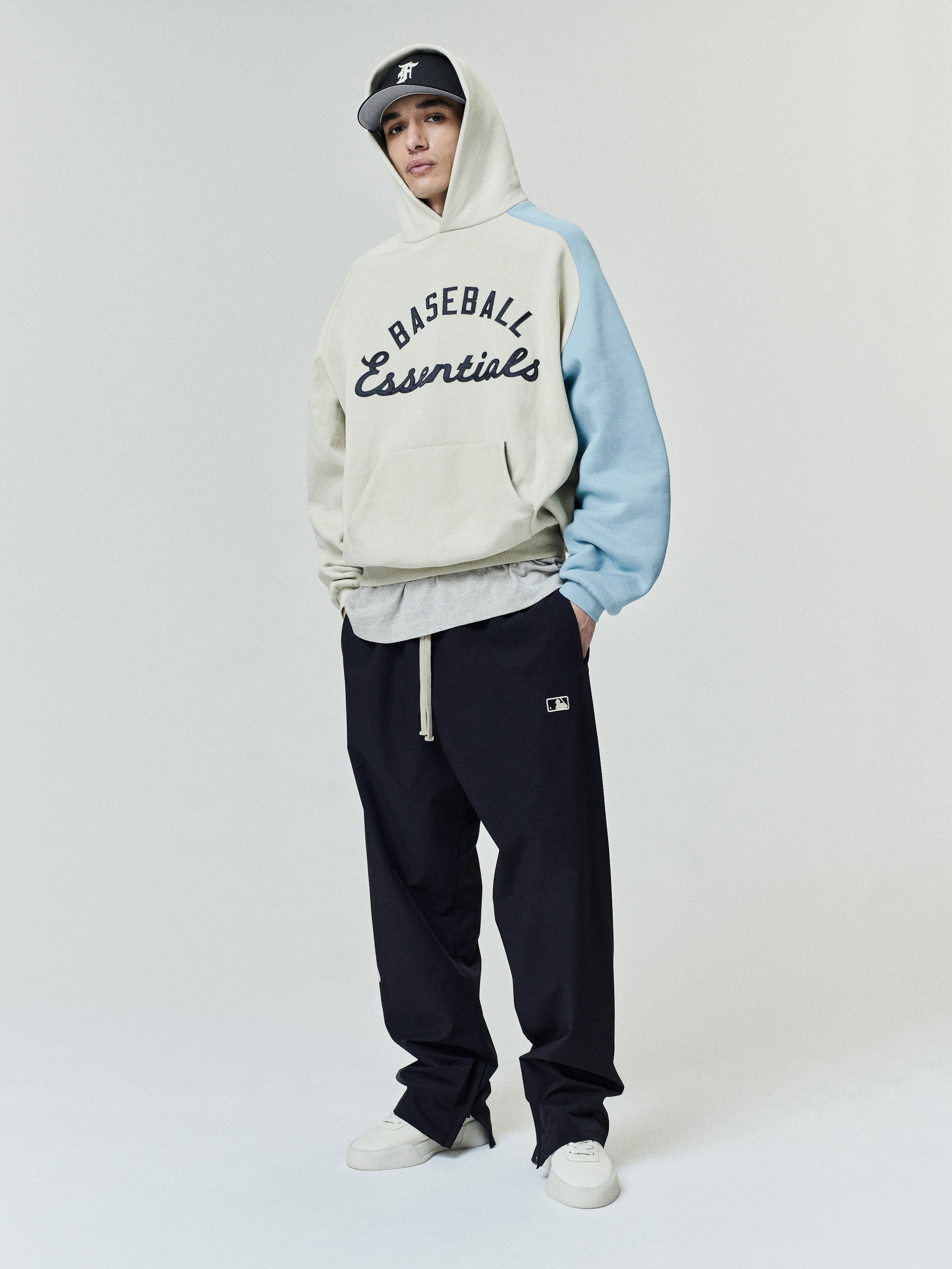 Model wearing a Mlb Raglan Sport Hoodie in cream with a contrasting light blue sleeve, featuring "Baseball Essentials" script across the chest, paired with relaxed black pants and a black cap.