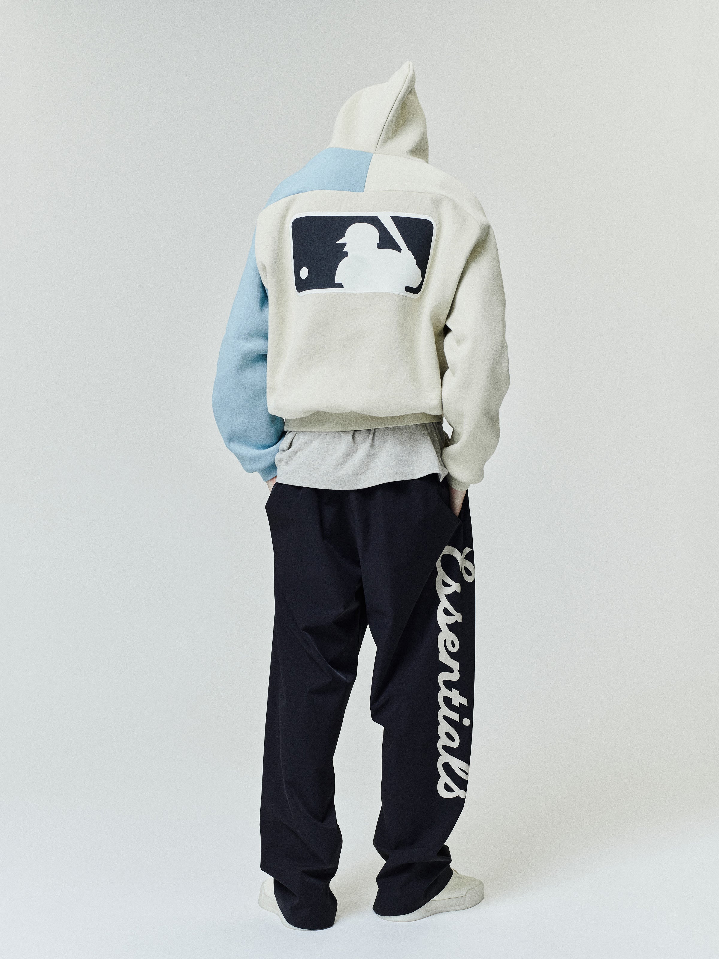 Model seen from the back wearing a Mlb Raglan Sport Hoodie in cream with a contrasting light blue sleeve, featuring a large baseball batter graphic on the back, paired with black pants with bold white lettering down the side for a casual outerwear look.