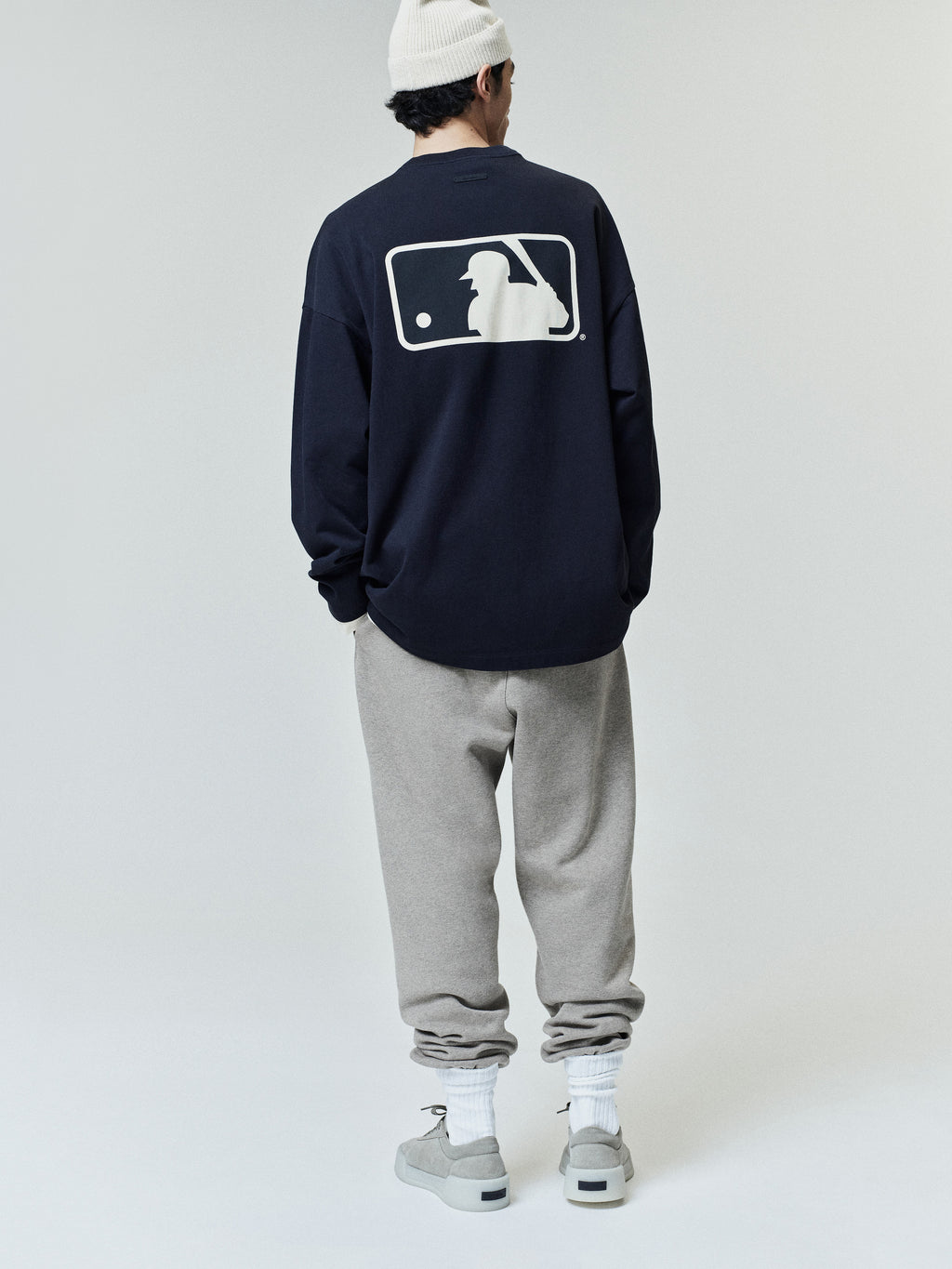 Rear view of a model wearing the MLB Boxy Long Sleeve Tee in navy blue with a large contrasting baseball silhouette logo on the back, paired with grey sweatpants and light sneakers.