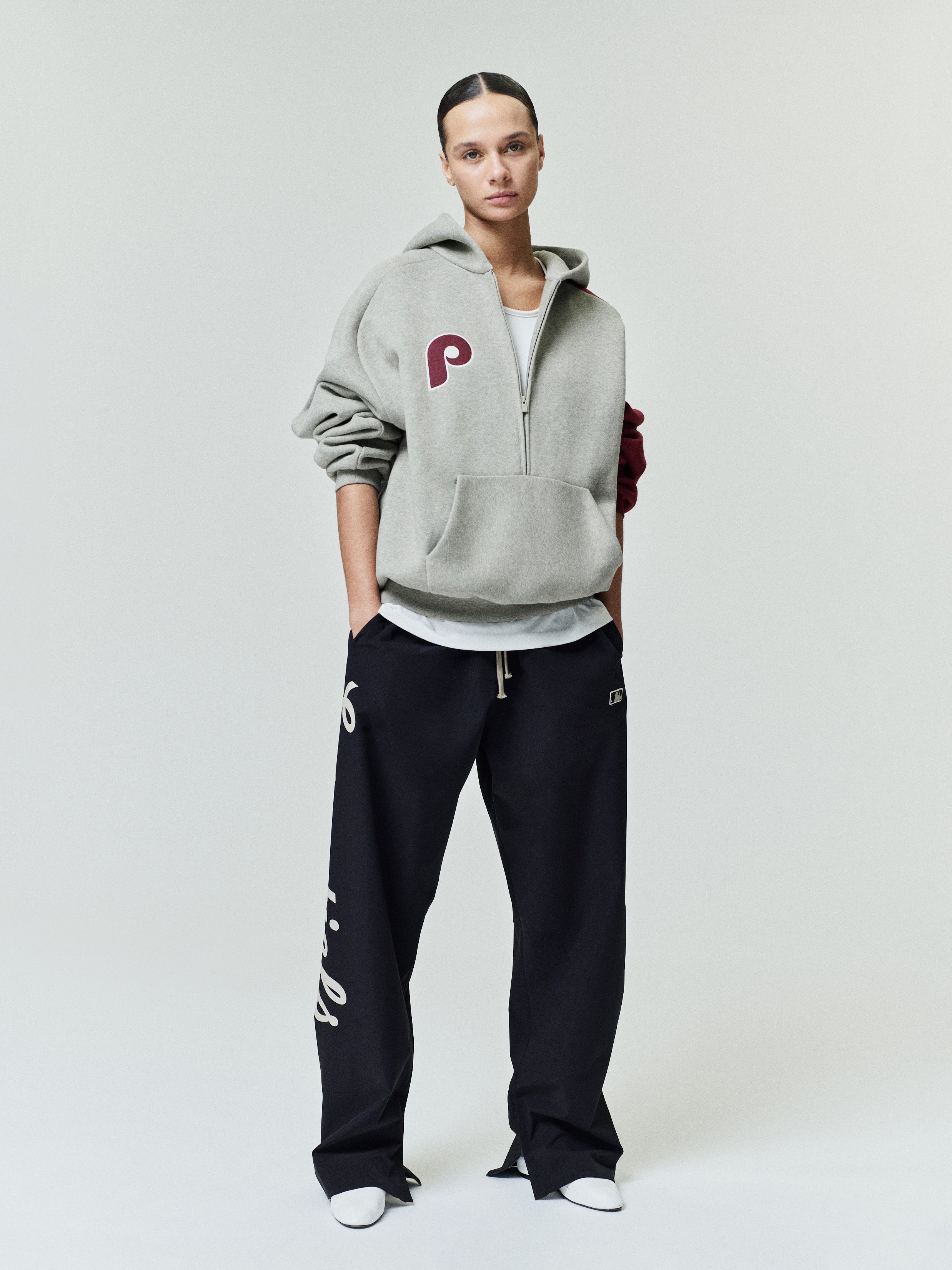 Model wearing a muted gray Phillies Raglan Half Zip Sport Hoodie with a maroon logo on the chest, paired with relaxed black pants featuring white text down the leg and white shoes.