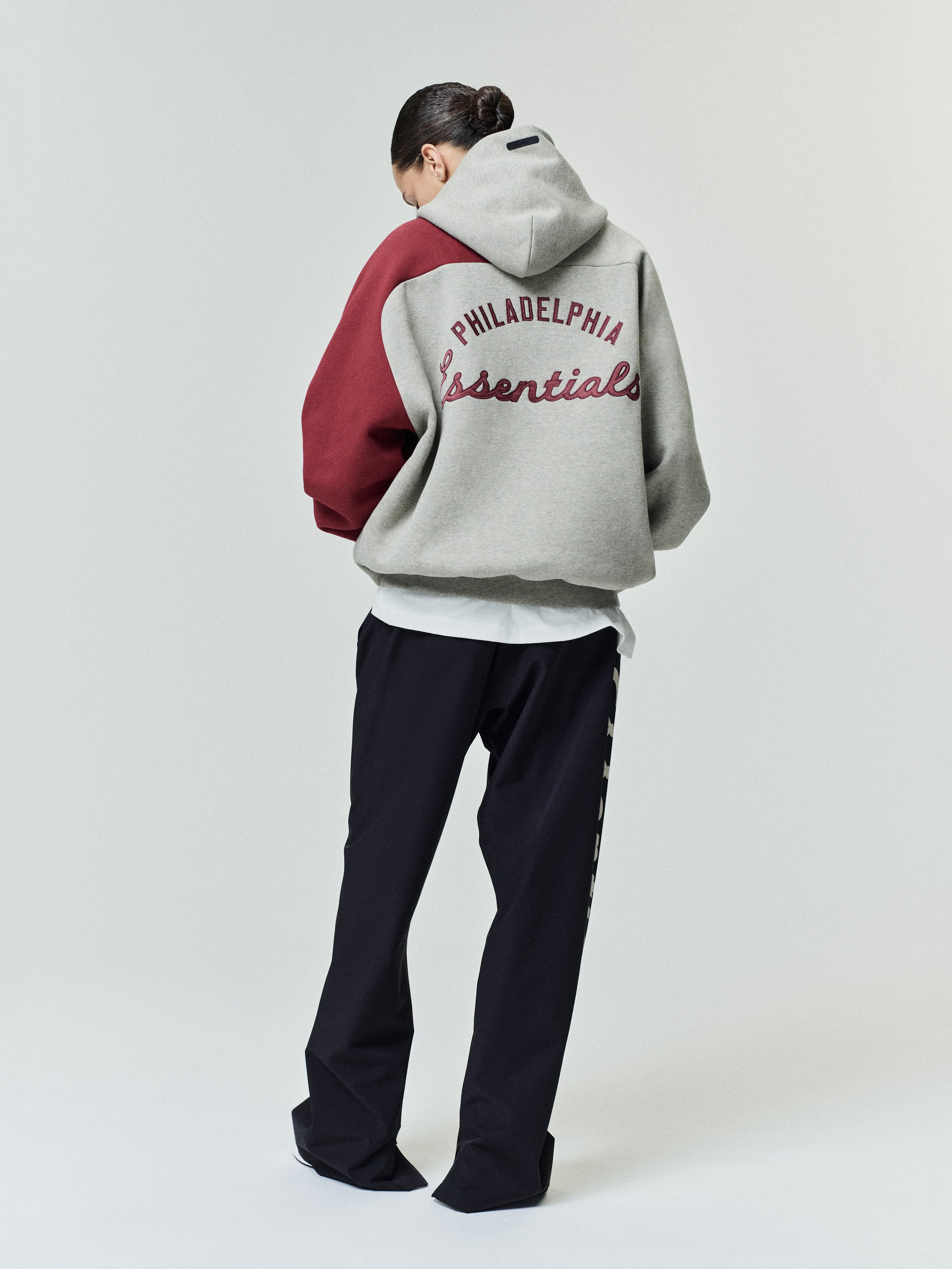 Model facing away wearing a Phillies Raglan Half Zip Sport Hoodie in light gray with a maroon sleeve and maroon "Philadelphia Essentials" text on the back, paired with black pants.