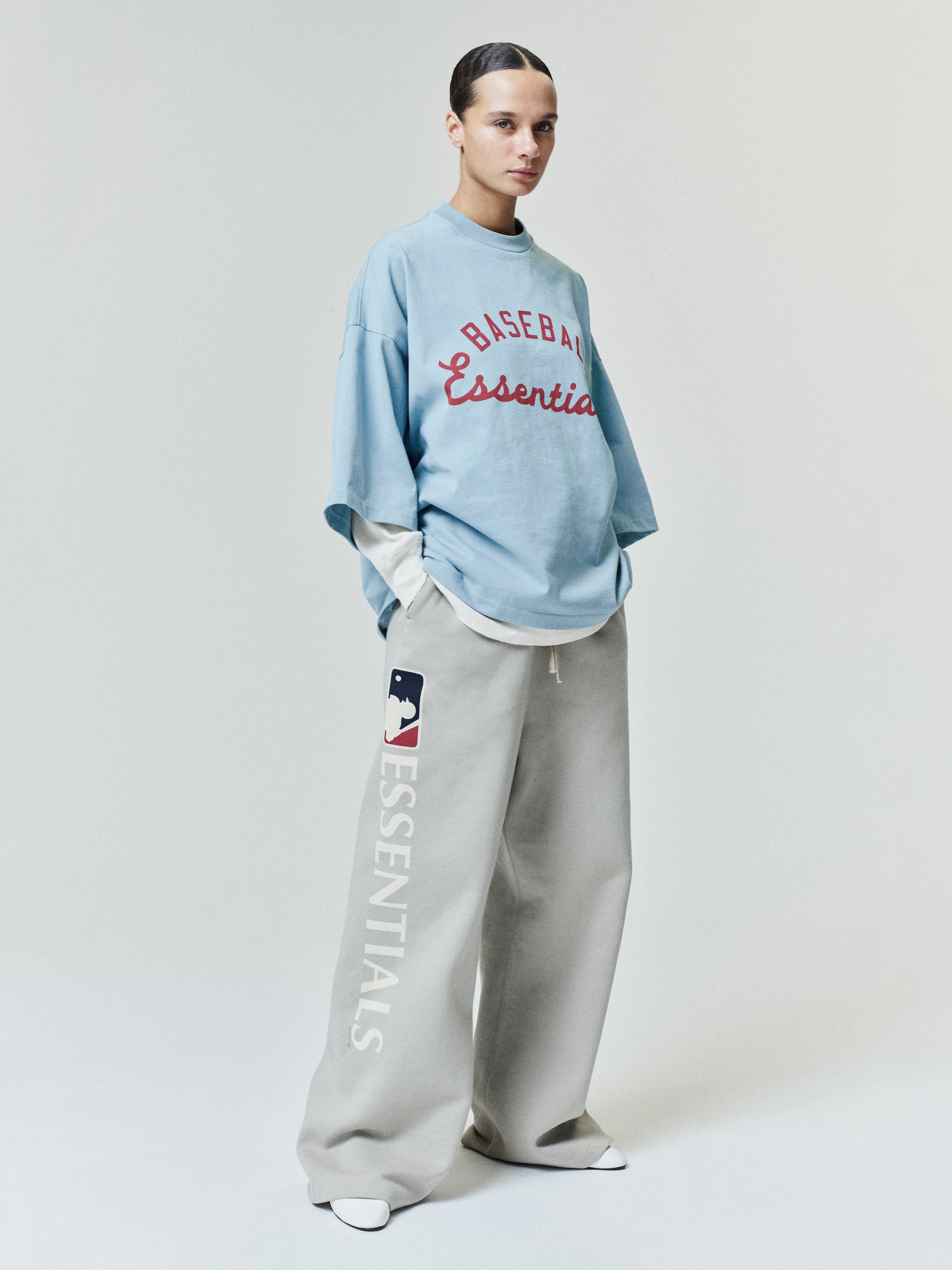 Model dressed in an oversized light blue Mlb Merch Tee layered over a long-sleeve shirt, paired with loose light beige sweatpants featuring vertical logo lettering and relaxed fit bottoms.