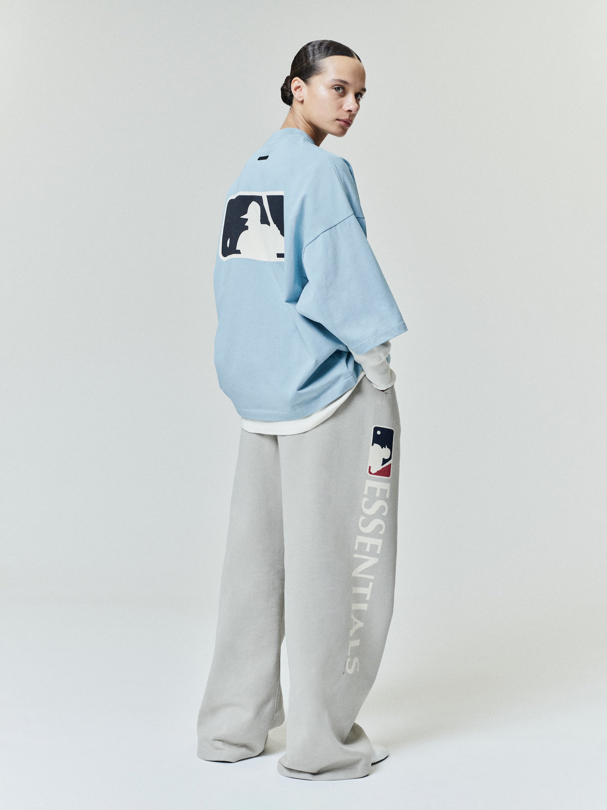 Model wearing a light blue Mlb Merch Tee with a large contrasting graphic on the back, paired with loose light gray sweatpants featuring vertical logo text and a small emblem near the pocket.