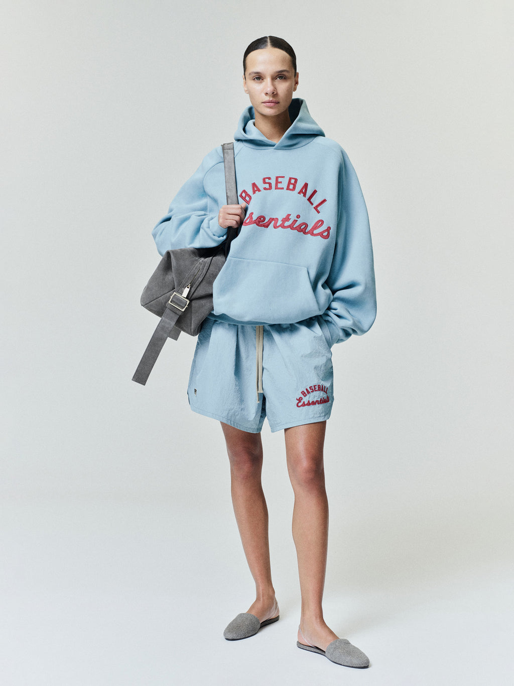 Model standing wearing a sky blue Mlb Raglan Sport Hoodie with red "Baseball Essentials" text on the chest, paired with matching shorts and carrying a grey suede shoulder bag in casual outerwear.