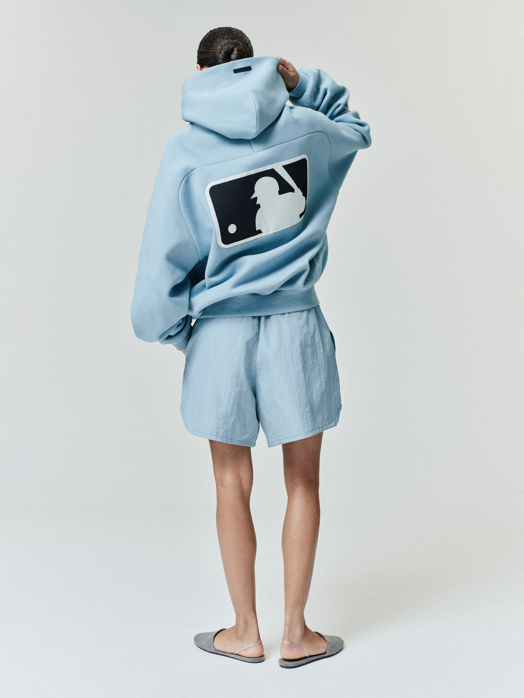 Rear view of a model wearing a sky blue Mlb Raglan Sport Hoodie featuring a large contrasting baseball batter silhouette graphic on the back, paired with matching shorts and casual slippers.