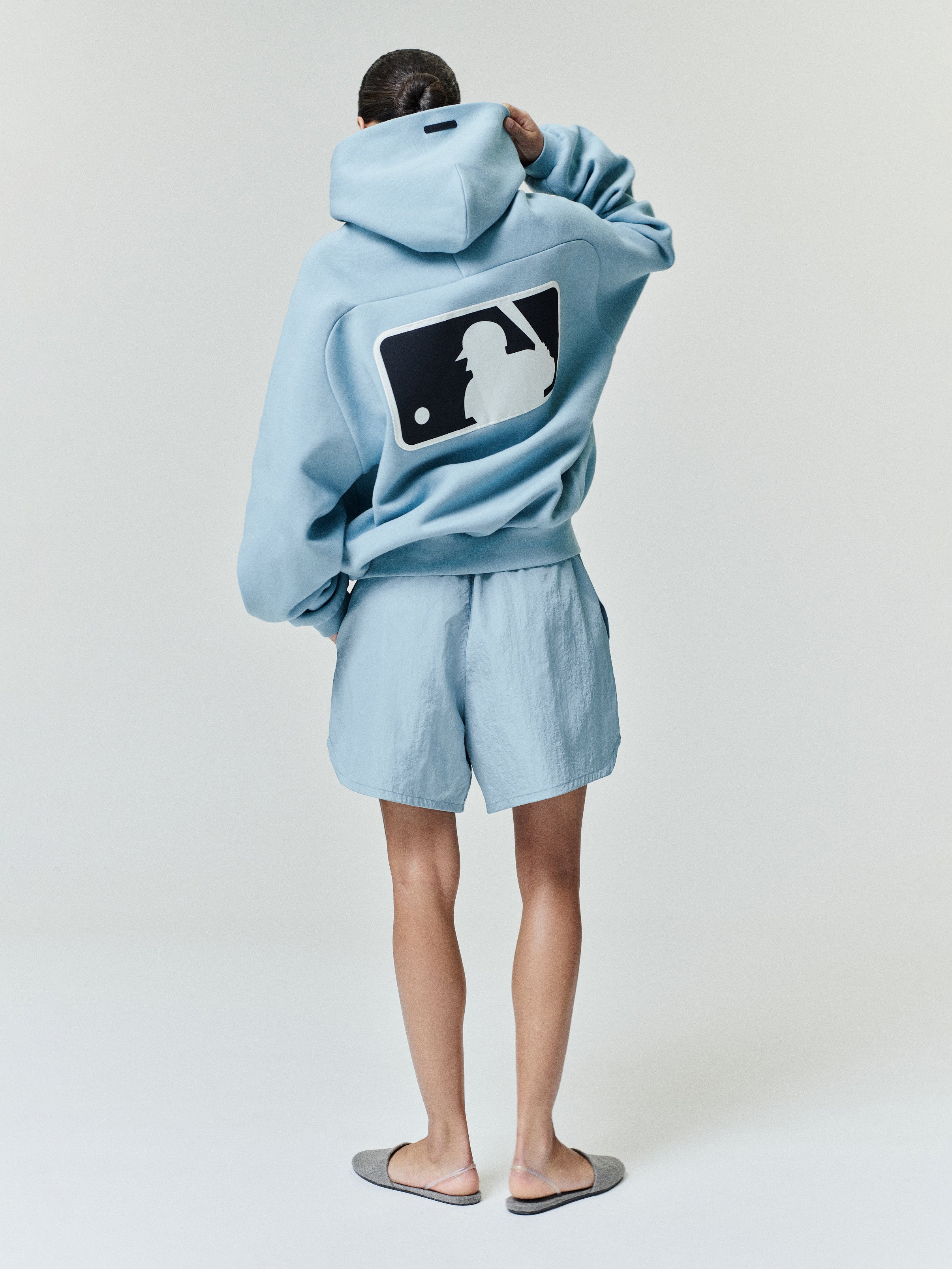 Rear view of a model wearing a sky blue Mlb Raglan Sport Hoodie featuring a large contrasting baseball batter silhouette graphic on the back, paired with matching shorts and casual slippers.