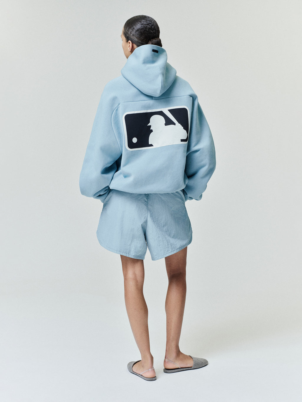 Rear view of a model wearing a sky blue Mlb Raglan Sport Hoodie with a large contrasting baseball batter logo centered on the back, paired with matching shorts and casual slippers.