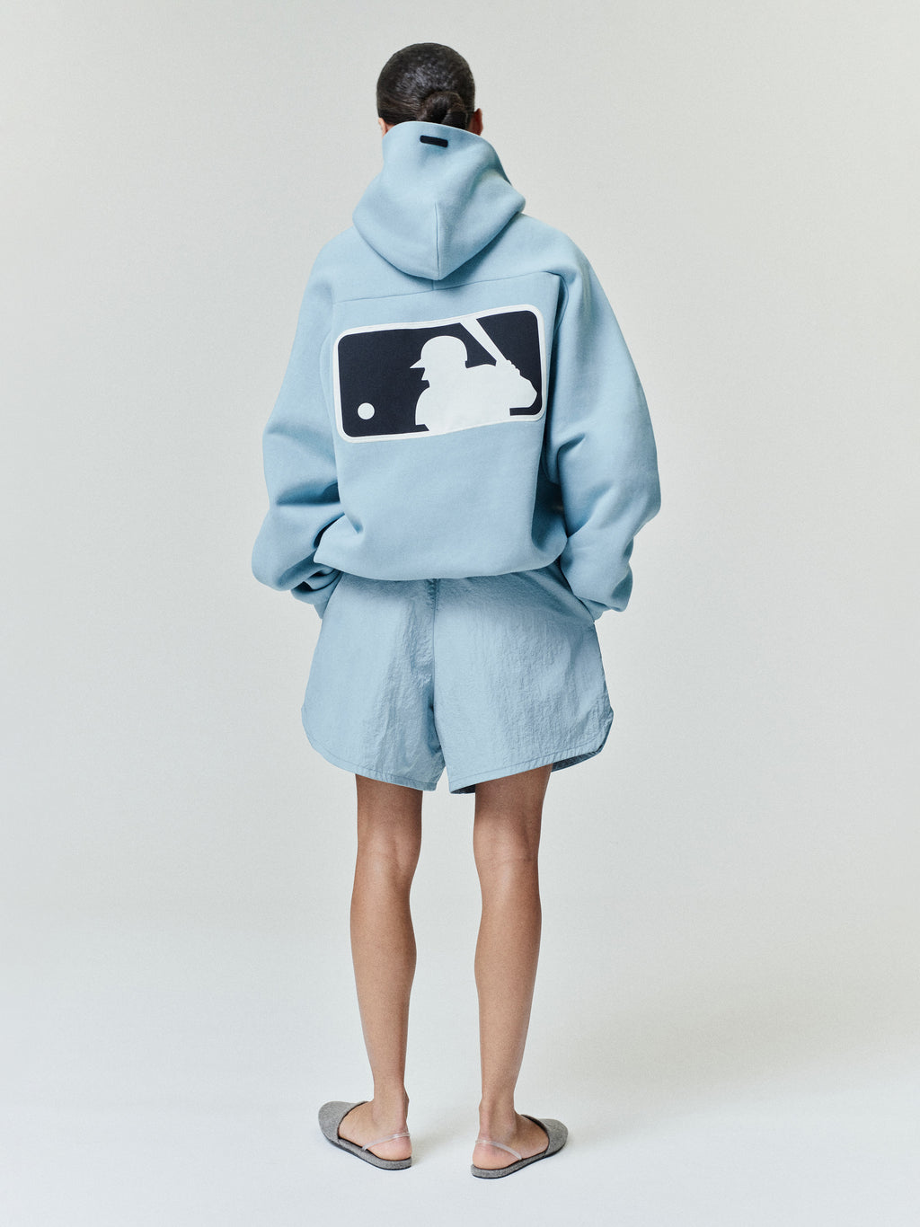 Model viewed from behind wearing a powder blue Mlb Raglan Sport Hoodie featuring a large contrasting baseball player graphic on the back, styled with matching relaxed shorts and slip-on footwear in casual outerwear.