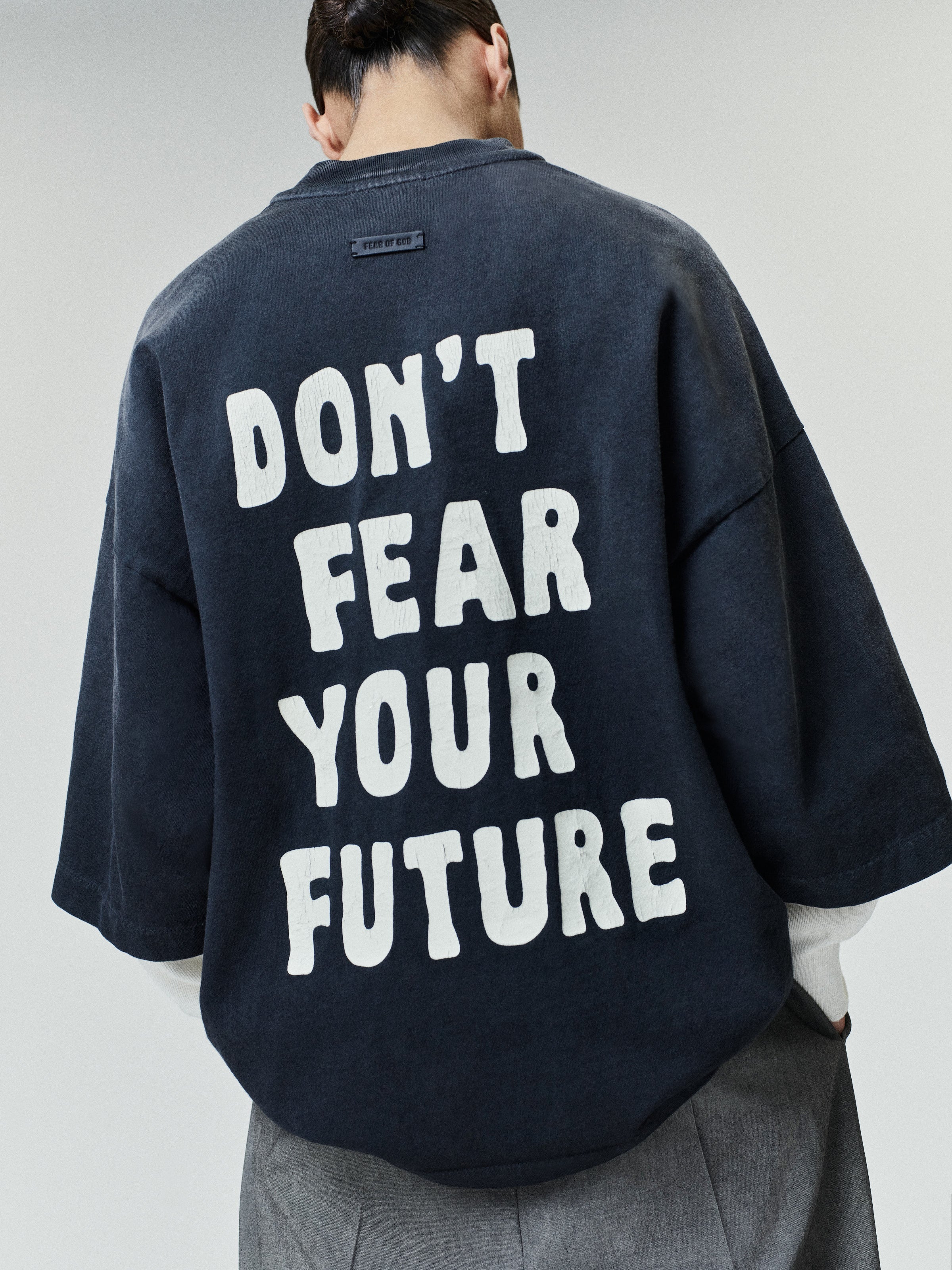 Person wearing an oversized dark washed t-shirt with large white block text on the back reading "DON'T FEAR YOUR FUTURE," paired with loose gray pants against a light background.