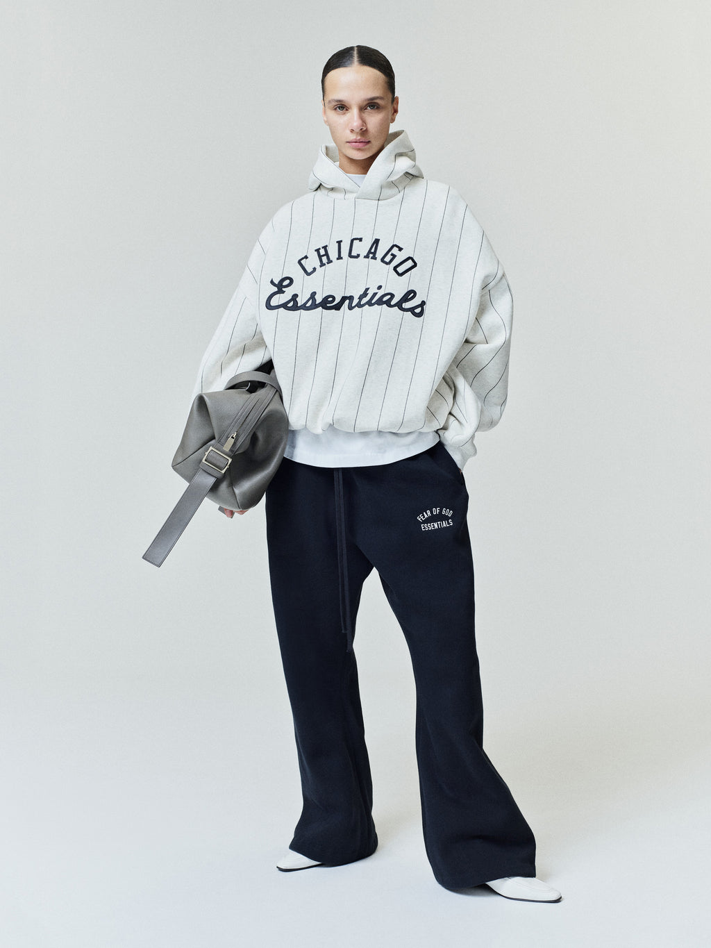 Model wearing White Sox Sport Hoodie in light gray with thin black vertical stripes and bold black "Chicago Essentials" text across the front, styled with relaxed black pants and carrying a gray bag.