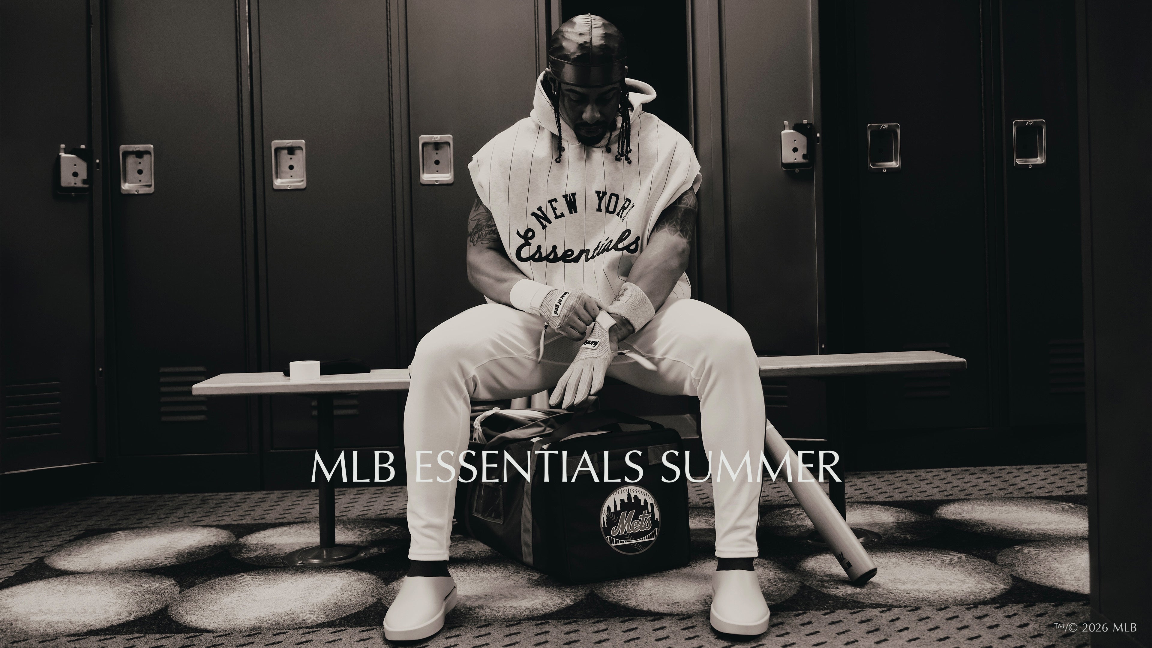 Man seated on a bench in a locker room wearing a sleeveless hoodie and sweatpants, holding a baseball bat with lockers in the background and soft overhead lighting.