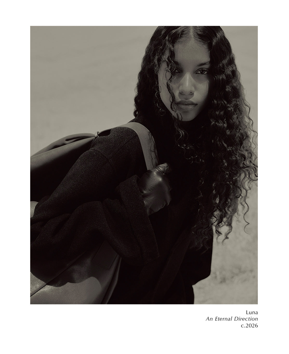 Monochrome portrait of a curly haired woman wearing a dark top and backpack while looking toward the camera against a softly textured neutral background with small text below