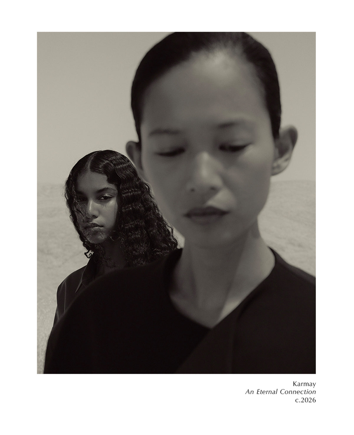 Monochrome layered portrait of two people in dark tops with the foreground figure looking downward and a curly haired woman behind them against a muted textured background with small text below