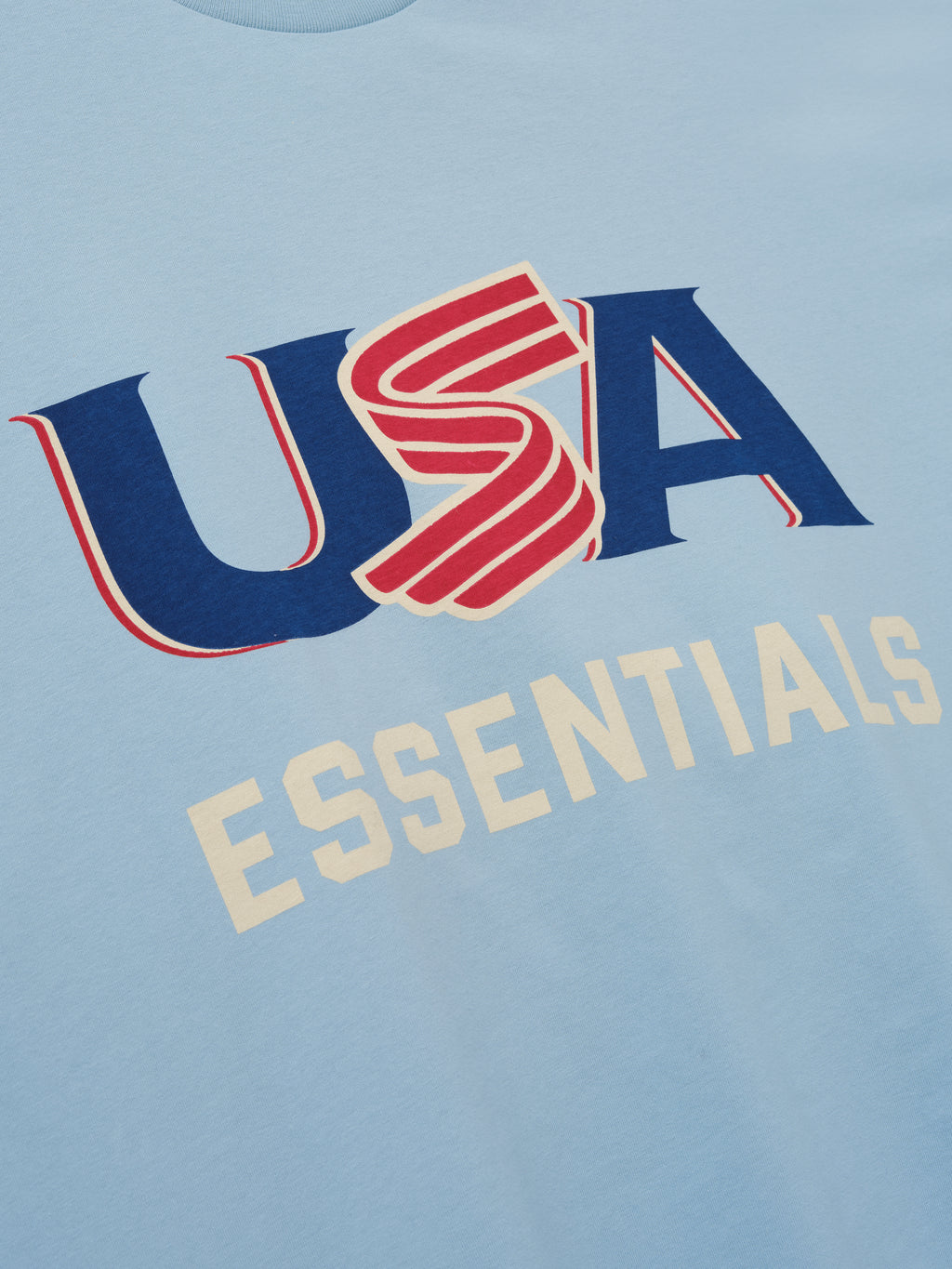 Close-up of light blue fabric with red, blue, and cream "USA" and cream "ESSENTIALS" text graphics on front center