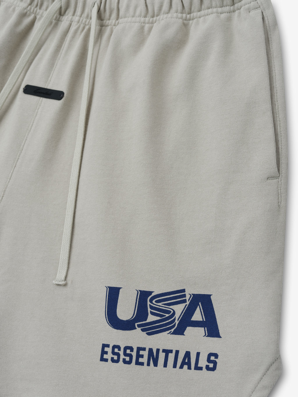Close-up of light beige sweatpants with elastic waistband, drawstrings, side pocket, and navy "USA ESSENTIALS" print on leg