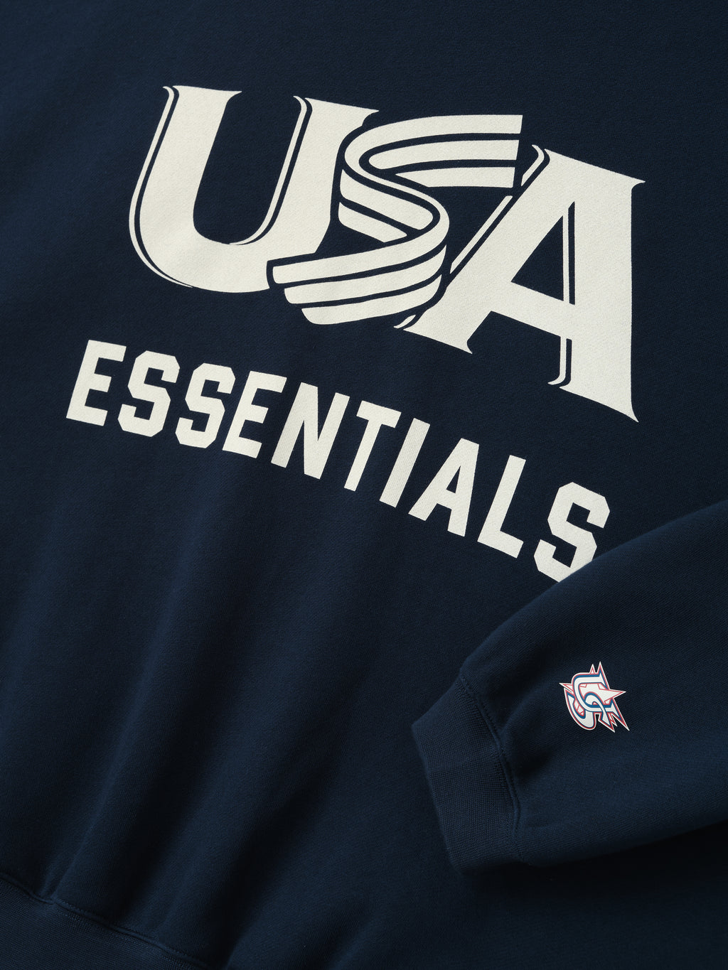 Close-up of navy sweatshirt with bold white "USA" graphic and "ESSENTIALS" text and small logo on sleeve cuff