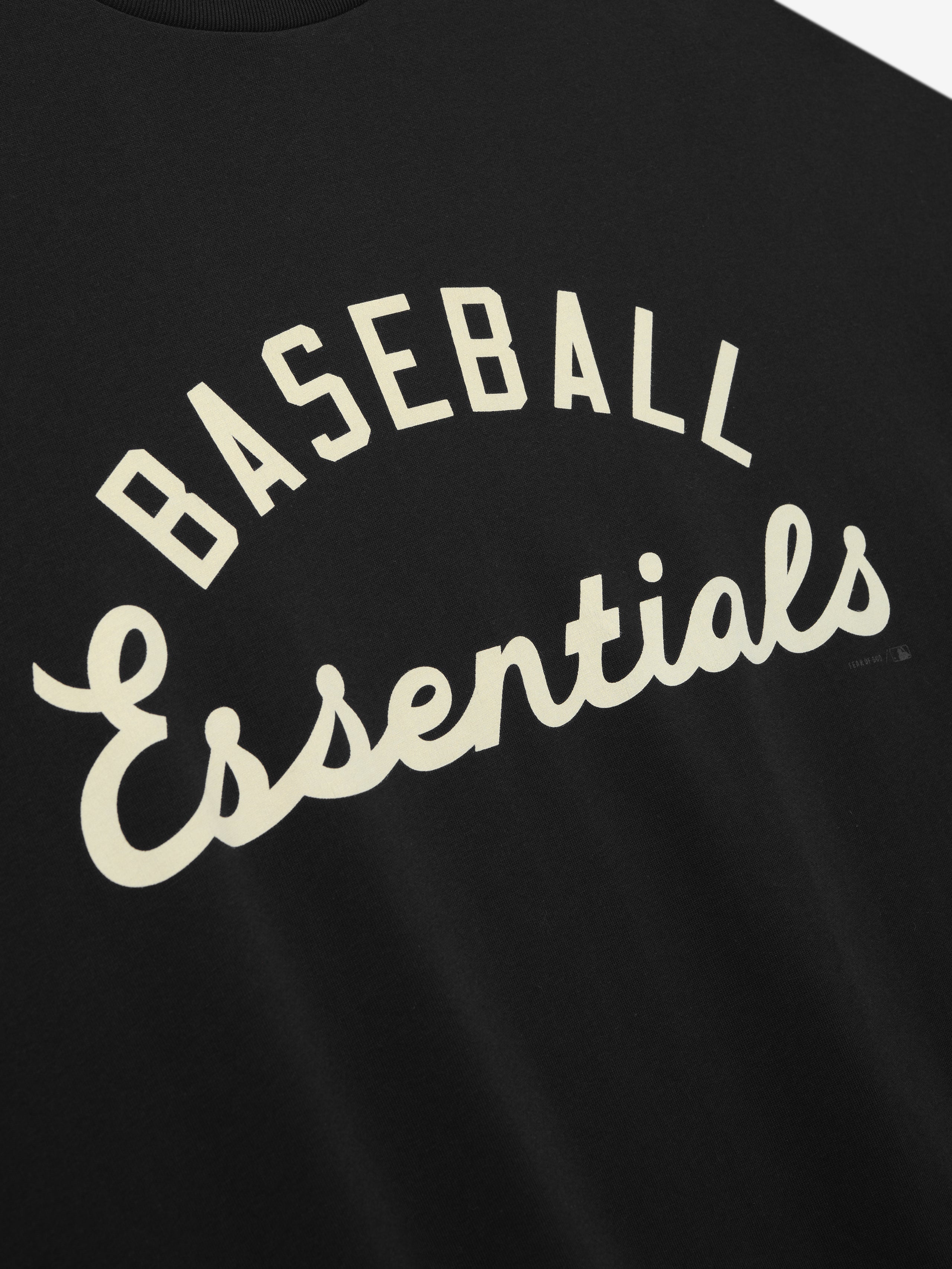 Close-up of black MLB Merch Tee fabric featuring a cream-colored "Baseball Essentials" curved script printed on soft cotton material, highlighting casual sporty style.