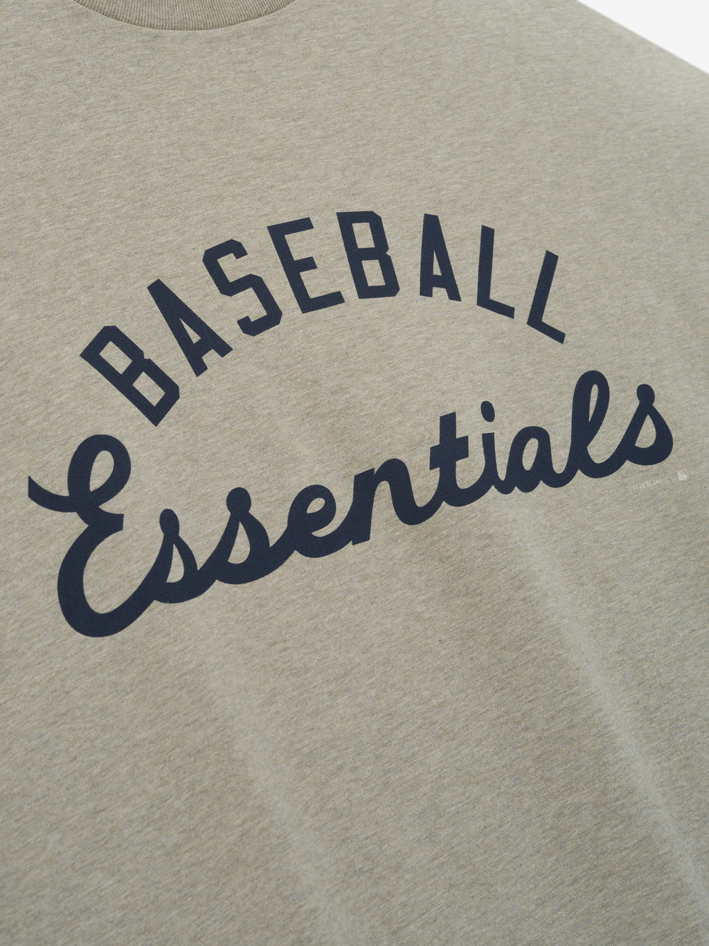 Close-up of a heathered beige Mlb Merch Tee fabric featuring bold navy blue script and block lettering that reads "BASEBALL Essentials" on a casual top.