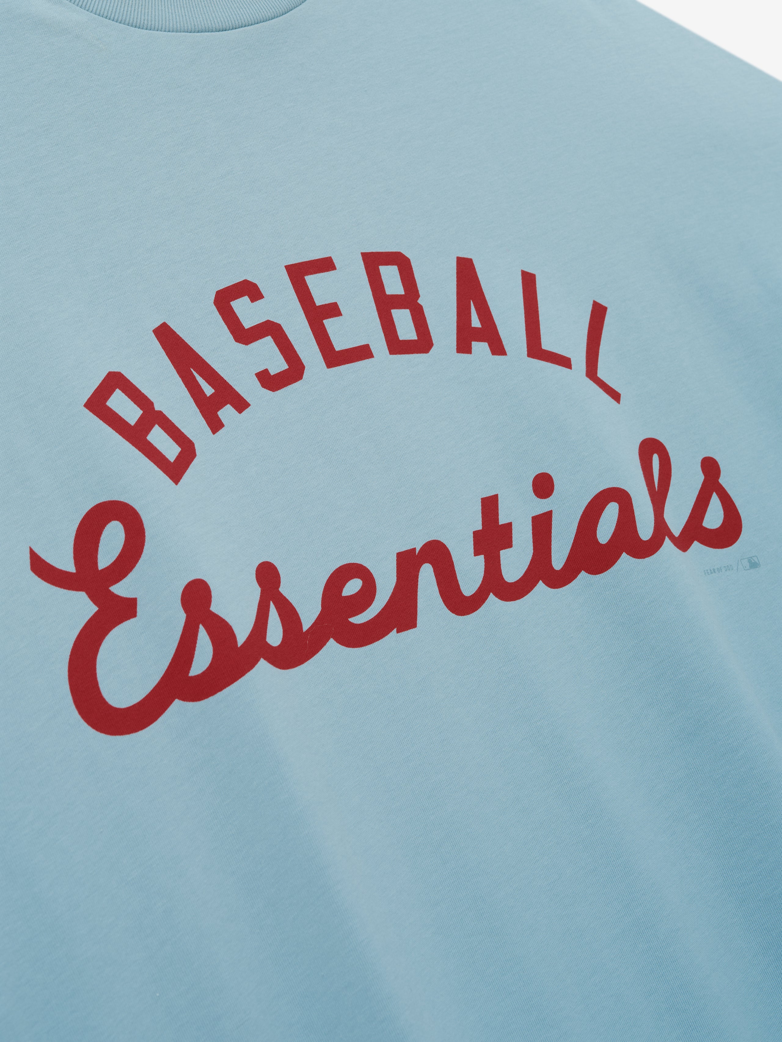 Close-up of light blue Mlb Merch Tee fabric featuring bold red lettering that reads "Baseball Essentials" in a mix of block and script fonts on a casual top.