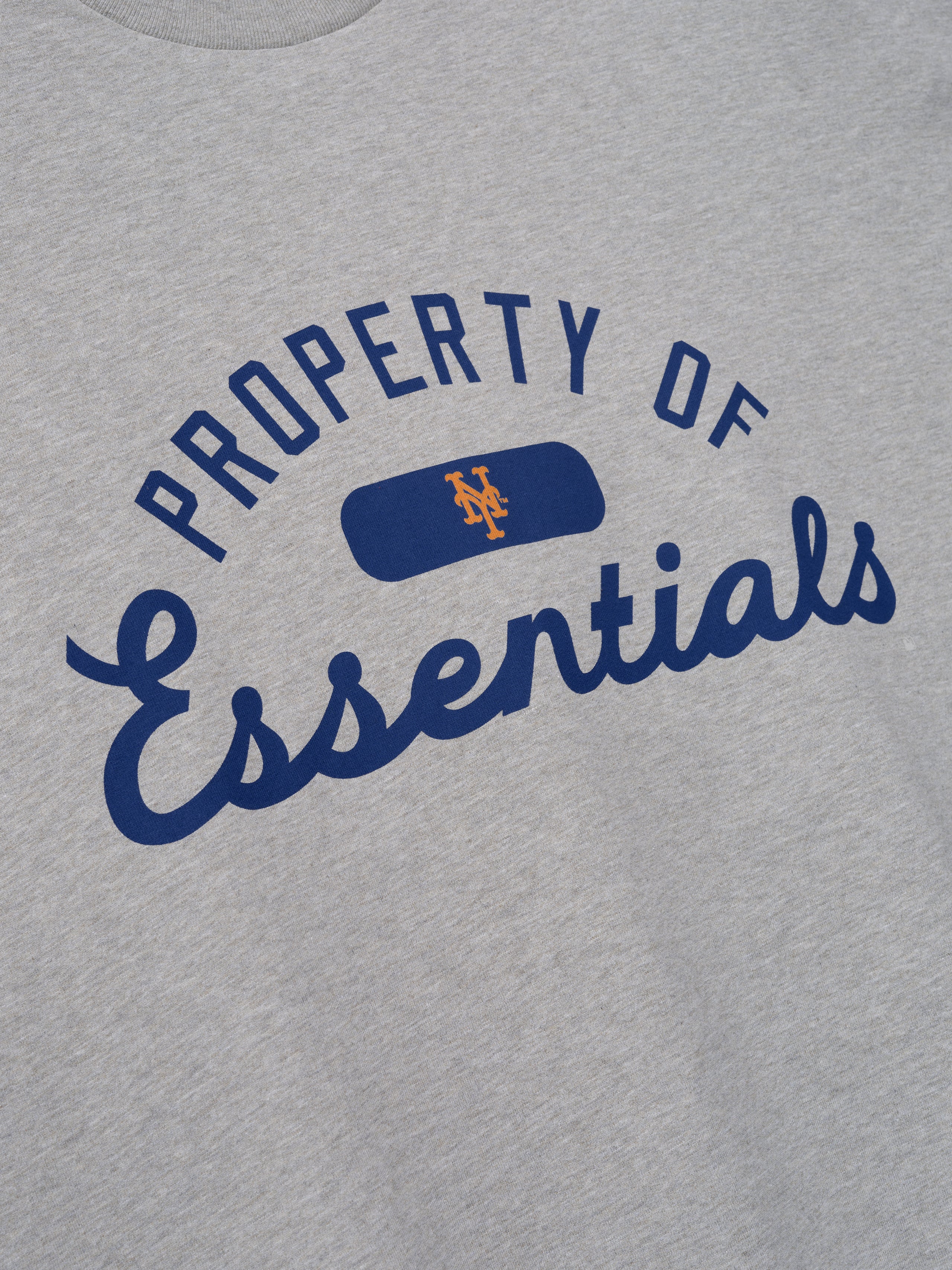 Close-up of the front of a Mets Merch Tee in light grey fabric featuring bold navy blue "Property of Essentials" text with a small orange Mets logo inside a blue patch, crafted from soft cotton.