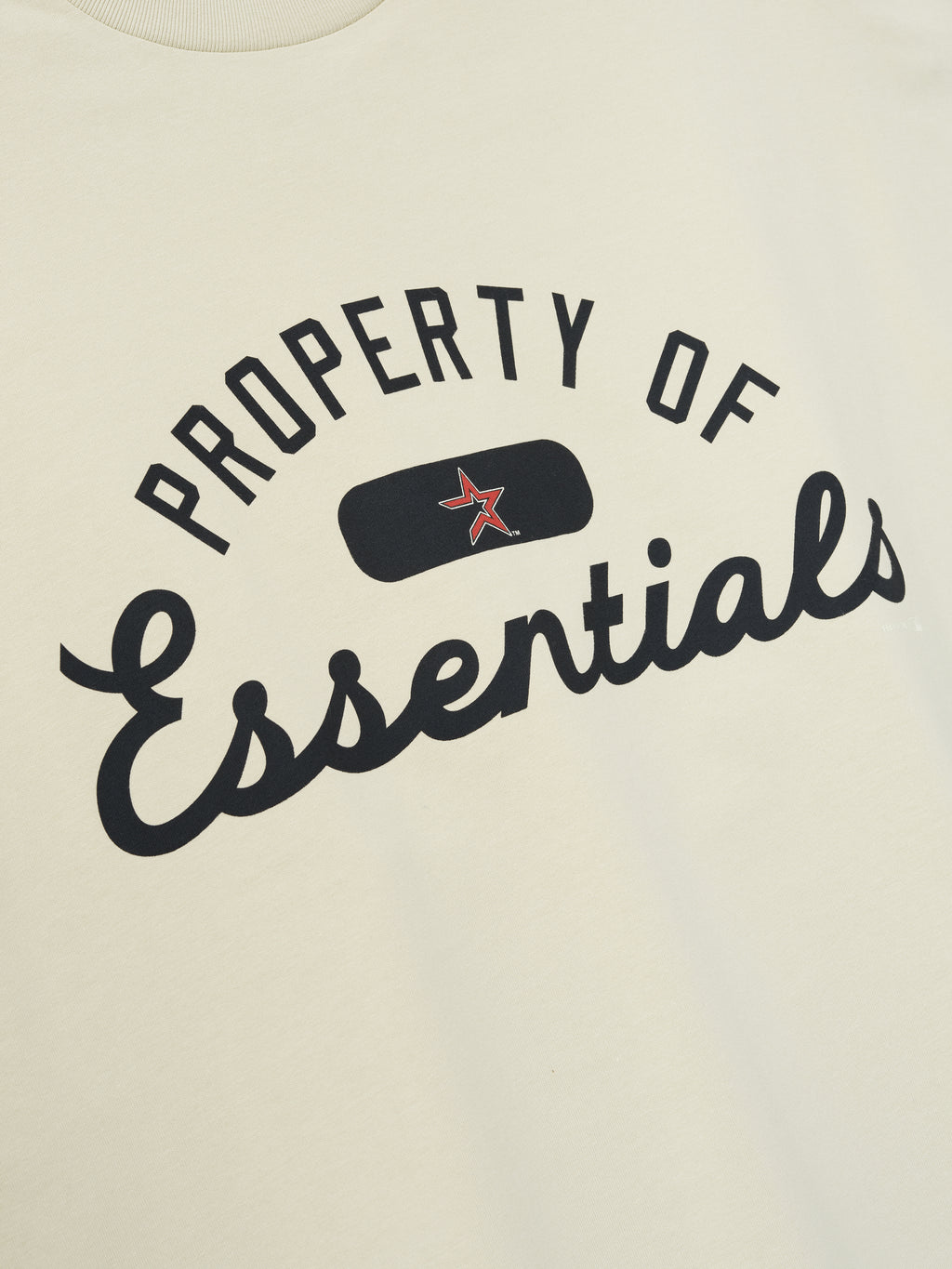 Close-up of Astros Merch Tee in a soft sandstone color featuring bold black "Property of Essentials" text with a small red star graphic on a smooth fabric surface.