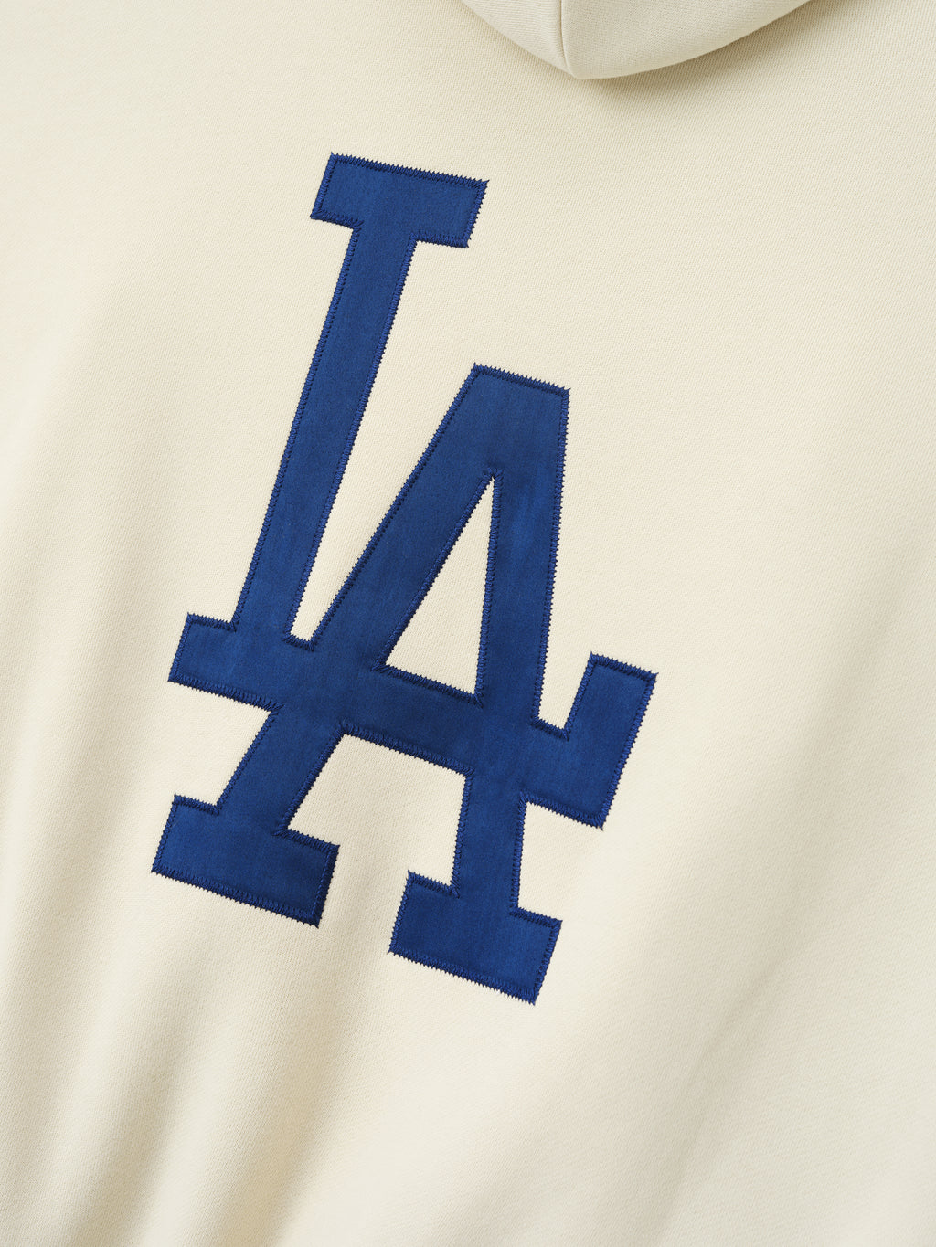 Close-up of cream fabric featuring a large navy blue stitched LA logo on the back of the Dodgers Sport Hoodie, showcasing detailed embroidery on soft outerwear material.