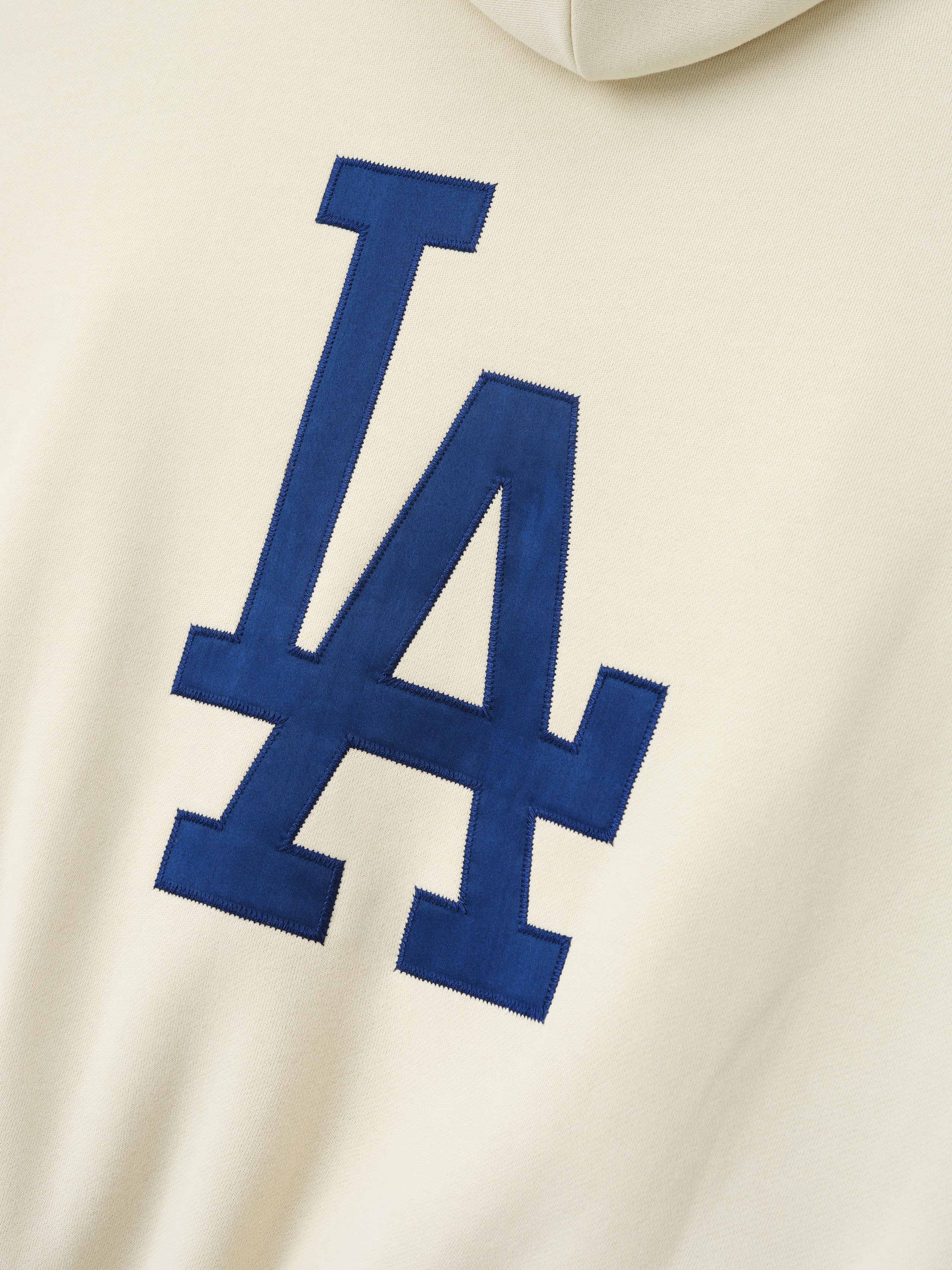Close-up of cream fabric featuring a large navy blue stitched LA logo on the back of the Dodgers Sport Hoodie, showcasing detailed embroidery on soft outerwear material.