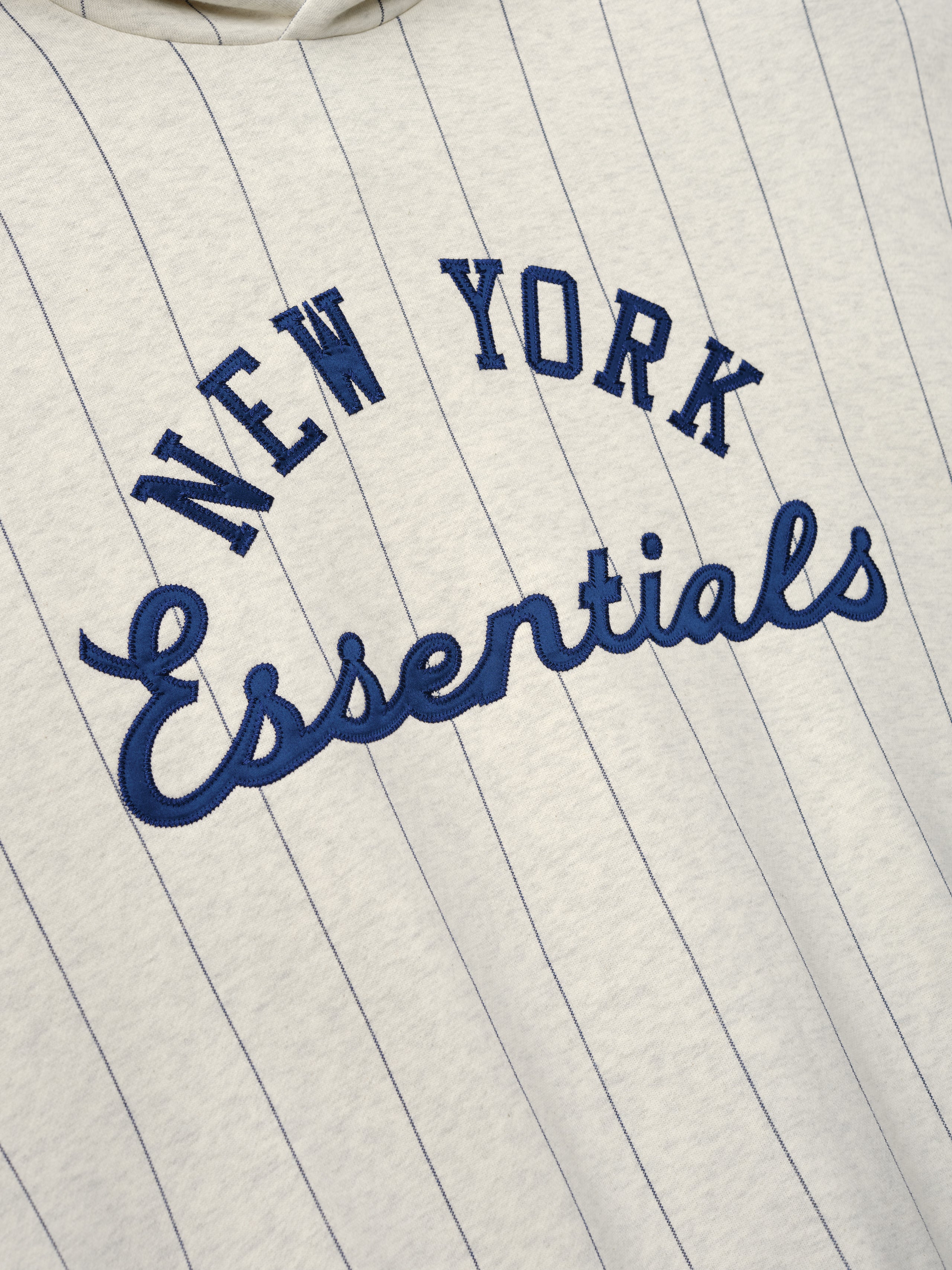 Close-up of the front of a Mets Sport Hoodie in oatmeal heather fabric featuring navy blue pinstripes and raised embroidered "New York Essentials" text on soft outerwear material.
