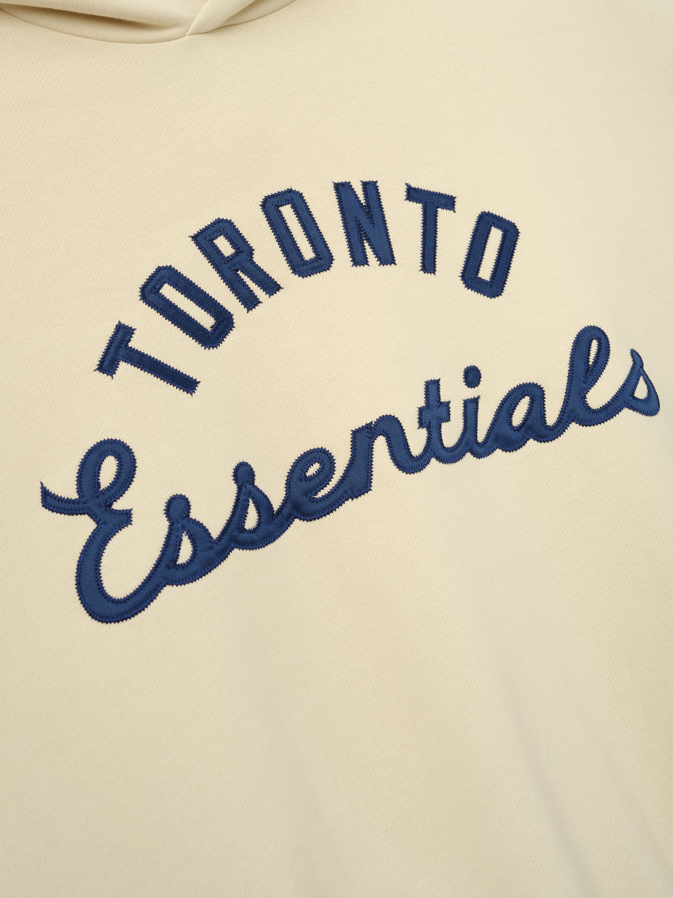 Blue Jays Sport Hoodie in a soft oatmeal tone featuring navy blue embroidered "Toronto Essentials" text in a stylish arch and script design on cozy outerwear fabric.