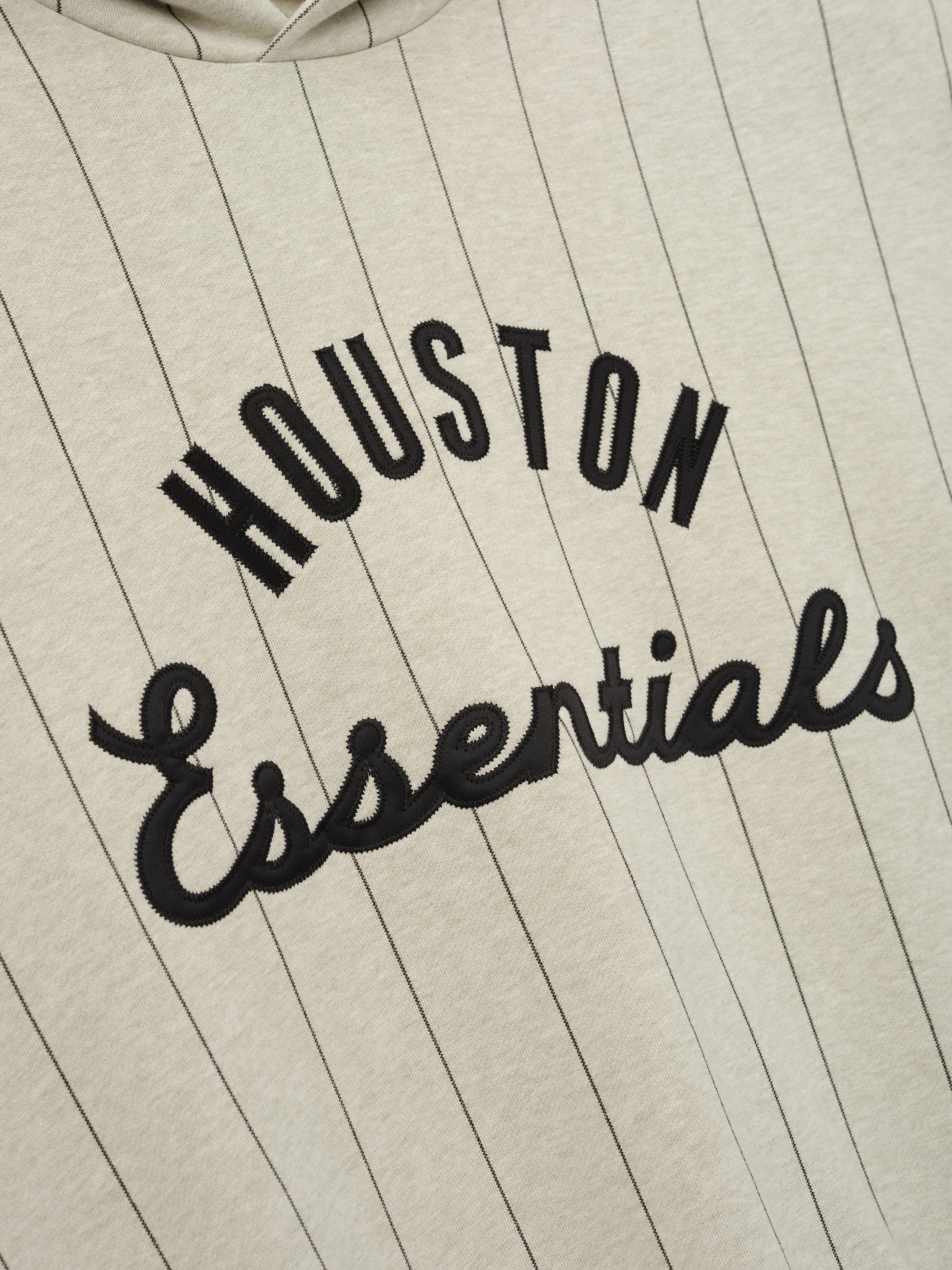 Close-up of Astros Sport Hoodie in a heather stone shade with thin black pinstripes, featuring bold black "HOUSTON Essentials" embroidered text on soft outerwear fabric.