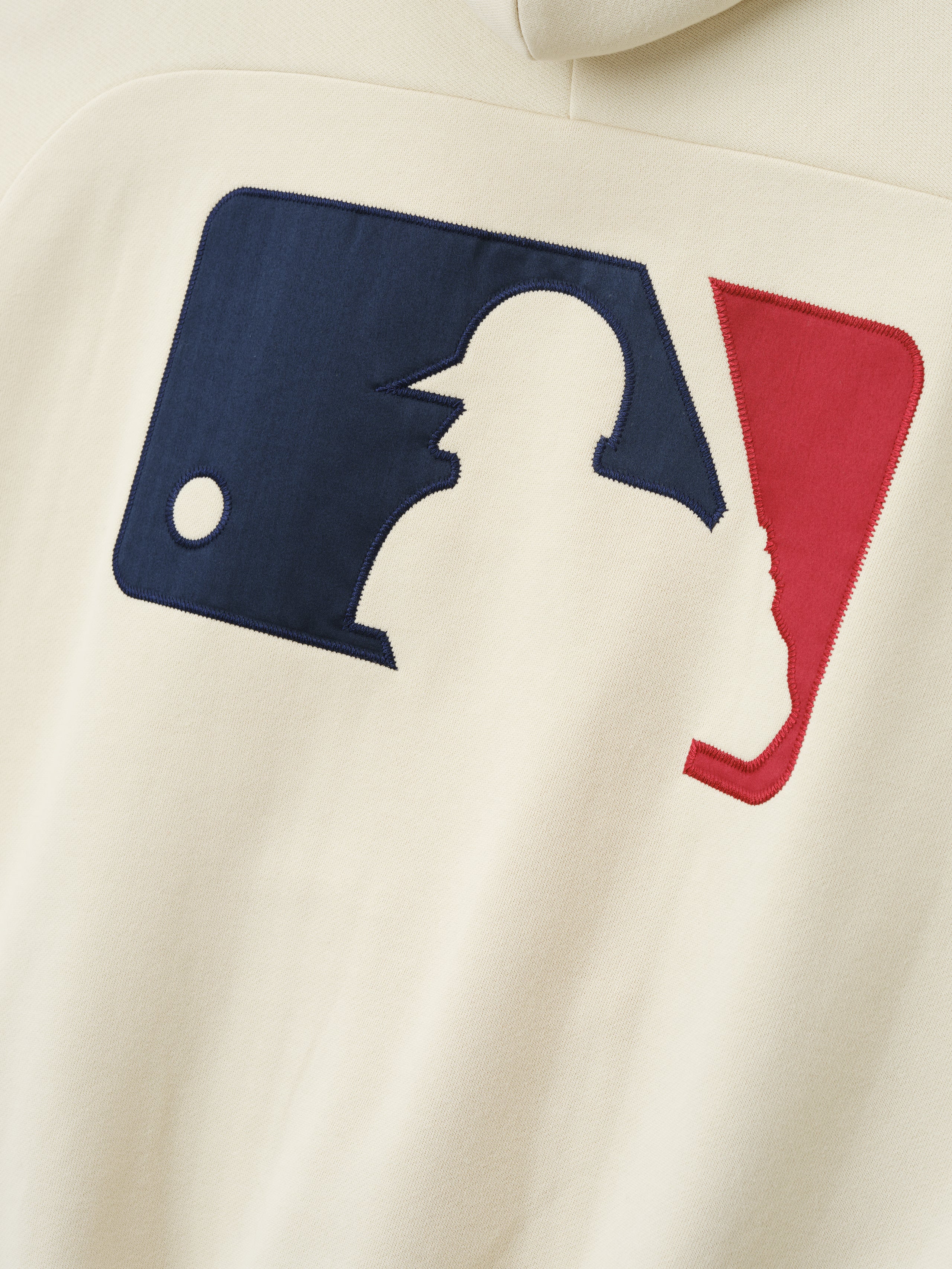 Close-up of the MLB Raglan Sport Hoodie in an off-white shade featuring a stitched navy and red baseball player silhouette emblem on soft outerwear fabric.