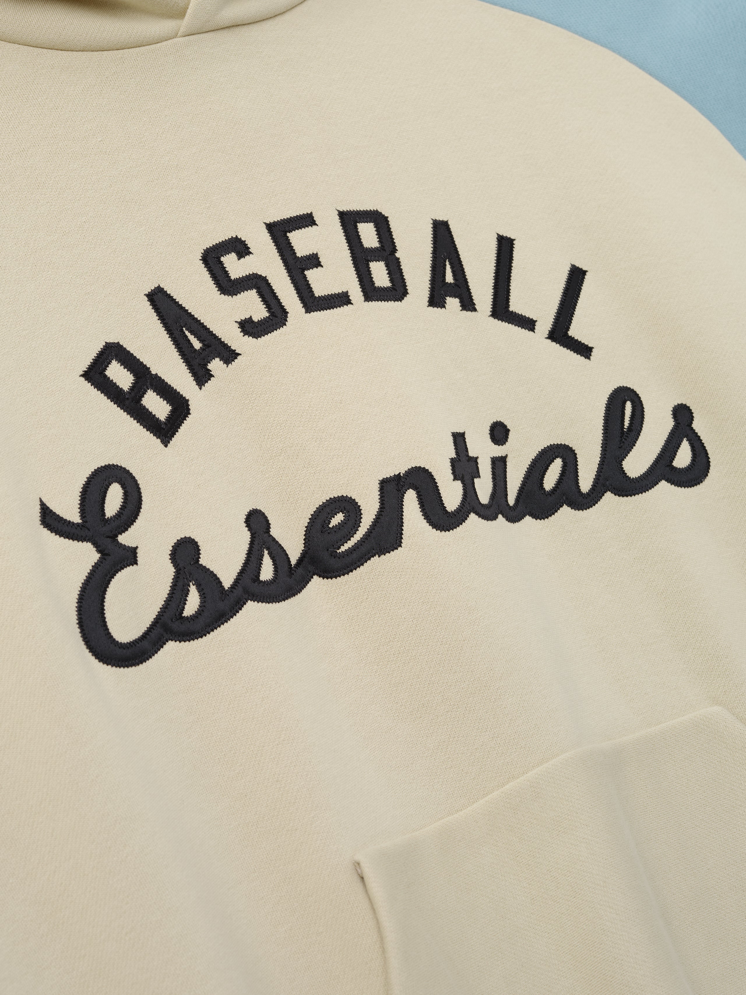 Close-up of the front of a Mlb Raglan Sport Hoodie in cream fabric with bold black embroidered "Baseball Essentials" text, highlighting soft material and detailed stitching on casual outerwear.