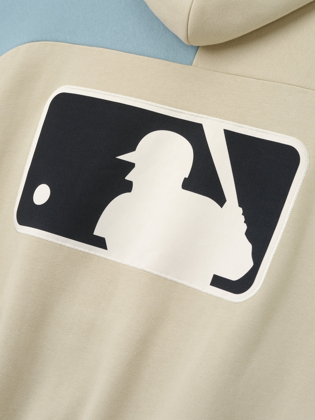 Close-up of the back of a Mlb Raglan Sport Hoodie in a soft sandstone color with a light blue sleeve, featuring a large contrasting silhouette graphic of a baseball batter and ball on comfortable outerwear.
