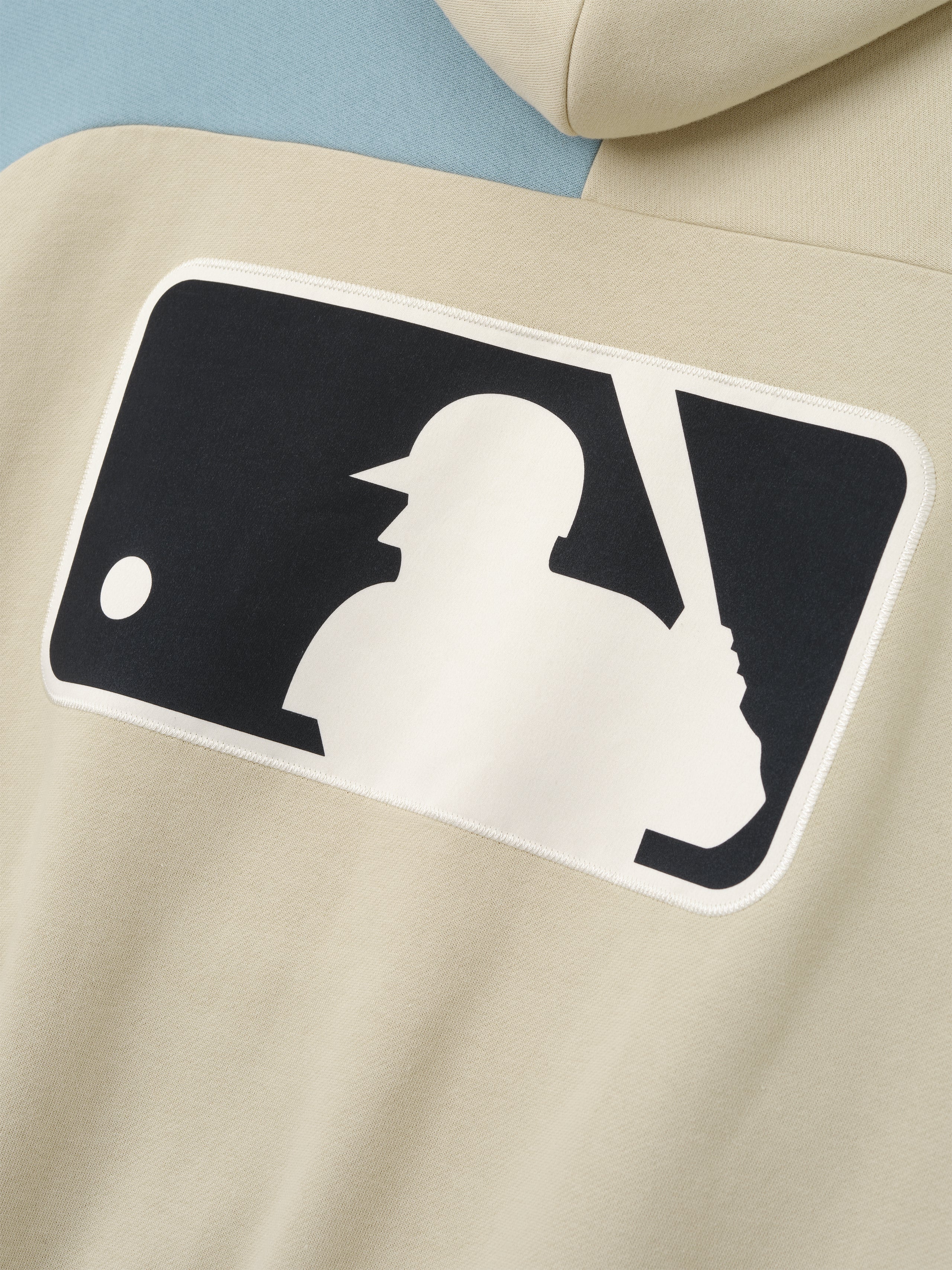 Close-up of the back of a Mlb Raglan Sport Hoodie in a soft sandstone color with a light blue sleeve, featuring a large contrasting silhouette graphic of a baseball batter and ball on comfortable outerwear.
