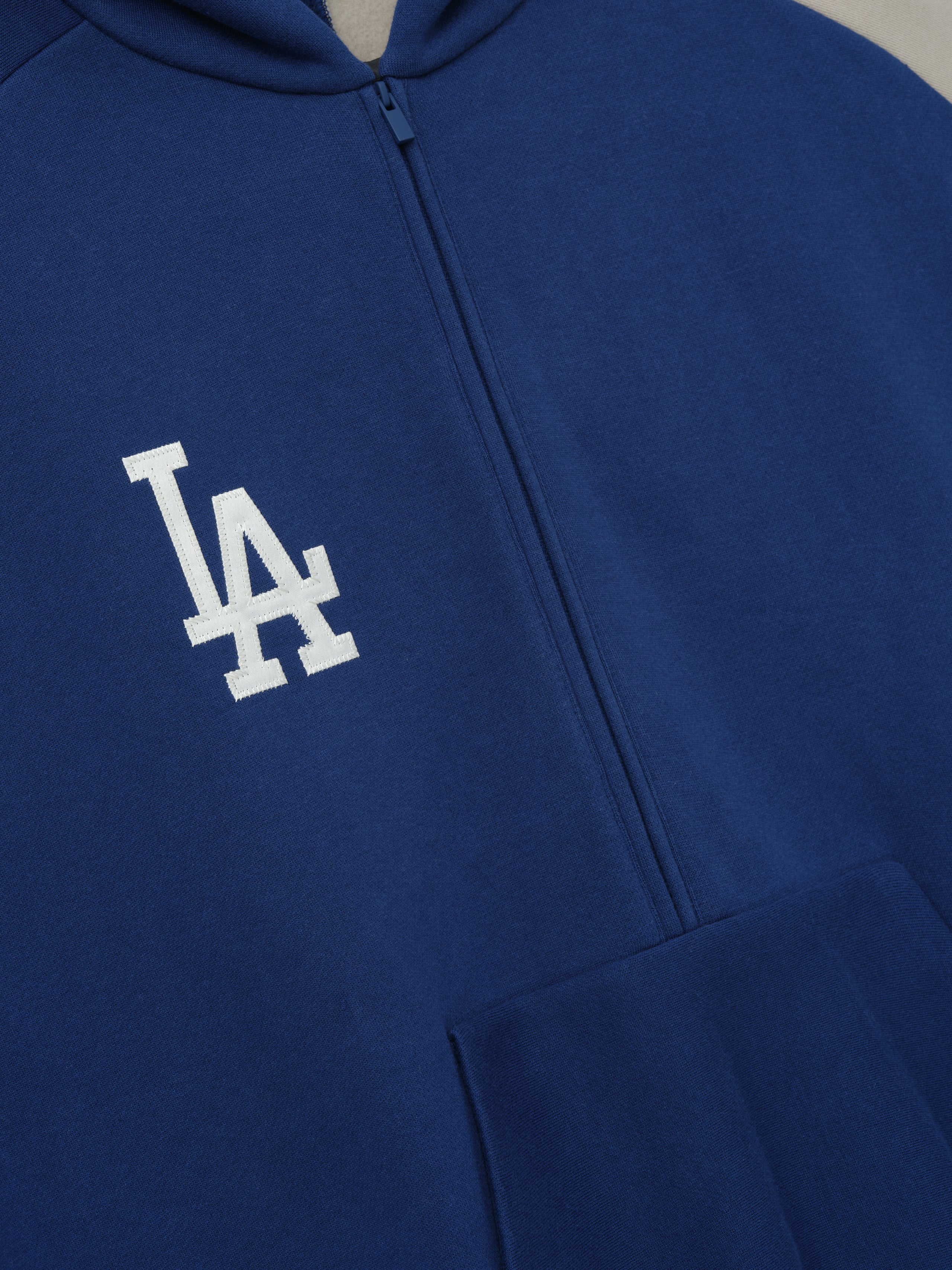 Close-up of Dodgers Raglan Half Zip Sport Hoodie in deep blue fabric featuring a white embroidered LA logo on the chest and a central zipper, styled as casual outerwear.