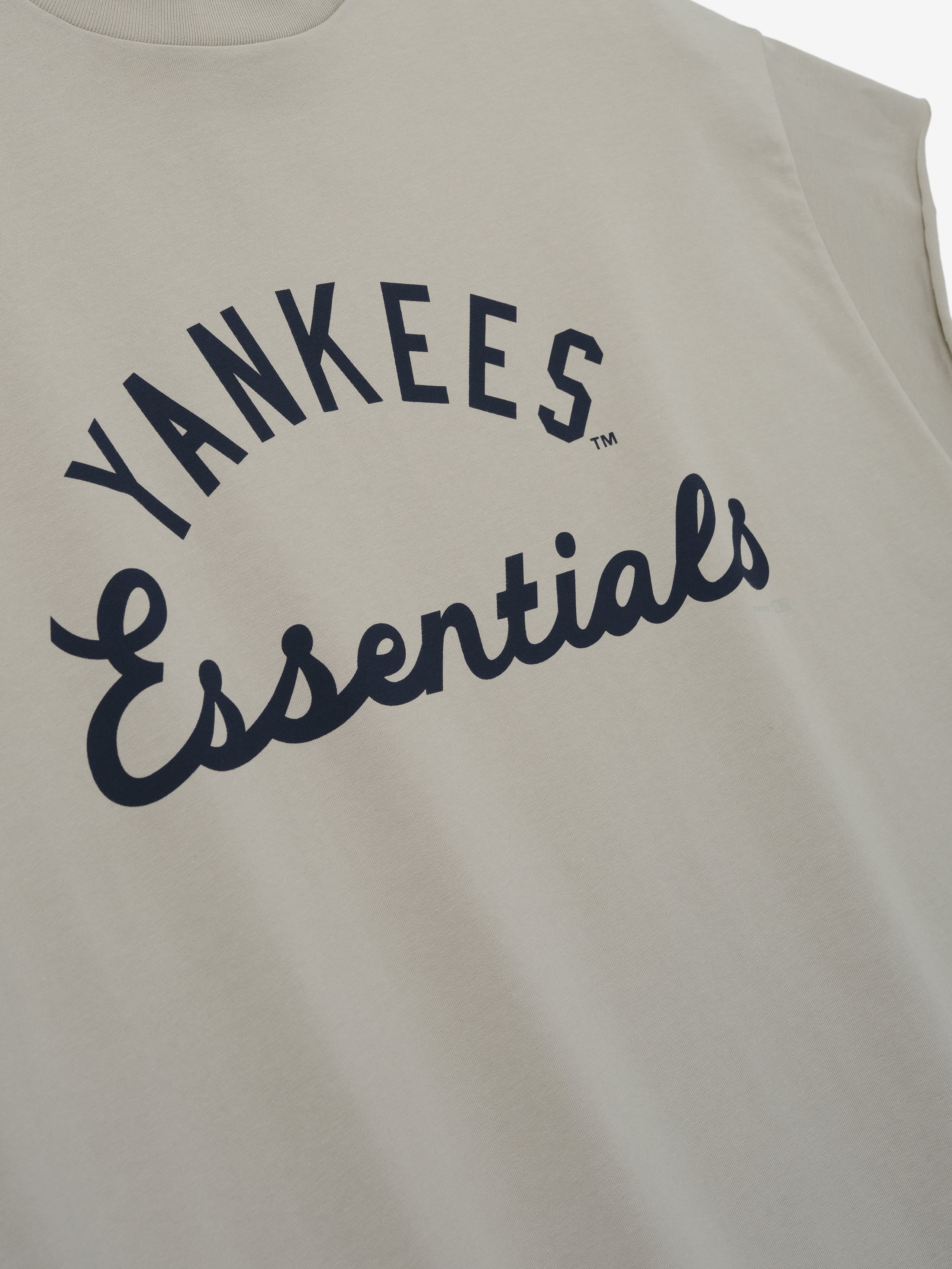 Yankees 90s Cut Off in a soft beige color featuring a sleeveless design with wide armholes and bold navy "Yankees Essentials" text printed across the chest on smooth cotton fabric.
