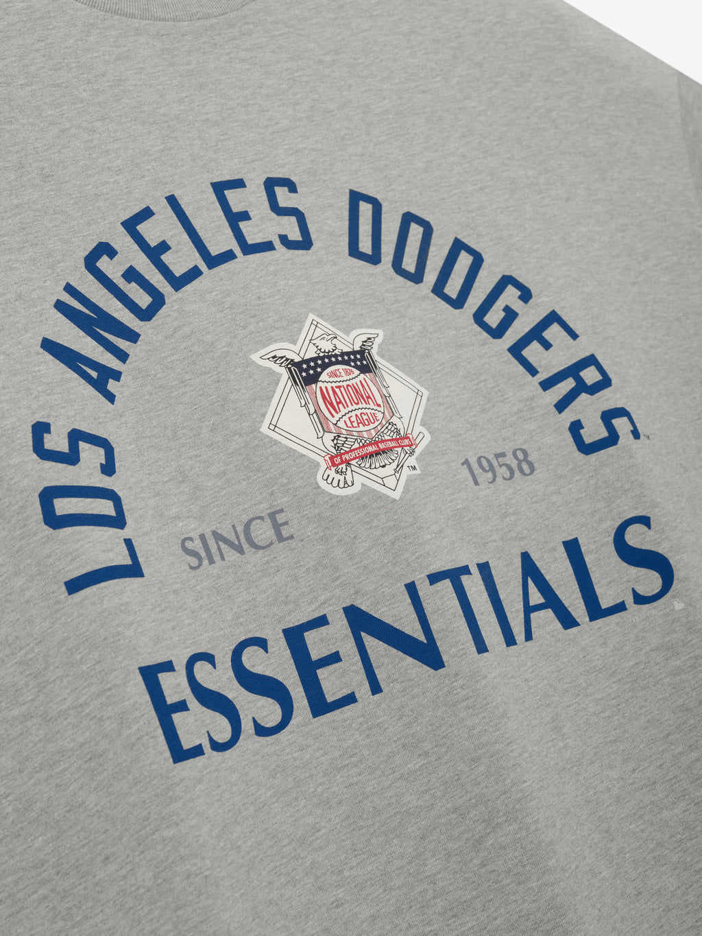 Close-up of the Dodgers NL Merch Tee in light gray featuring navy blue "Los Angeles Dodgers Essentials" text arched around a vintage-style National League emblem on soft fabric.