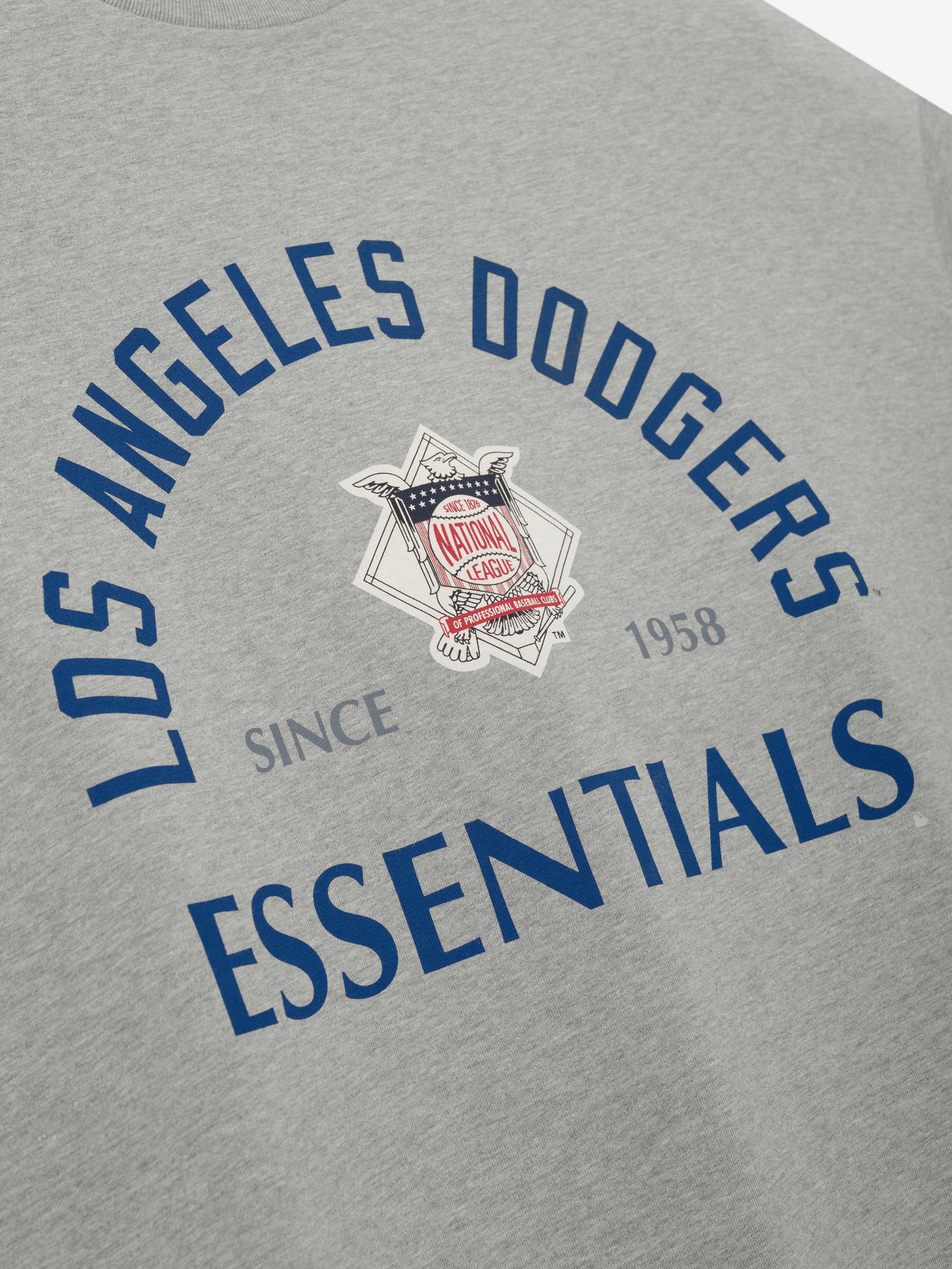 Close-up of the Dodgers NL Merch Tee in light gray featuring navy blue "Los Angeles Dodgers Essentials" text arched around a vintage-style National League emblem on soft fabric.
