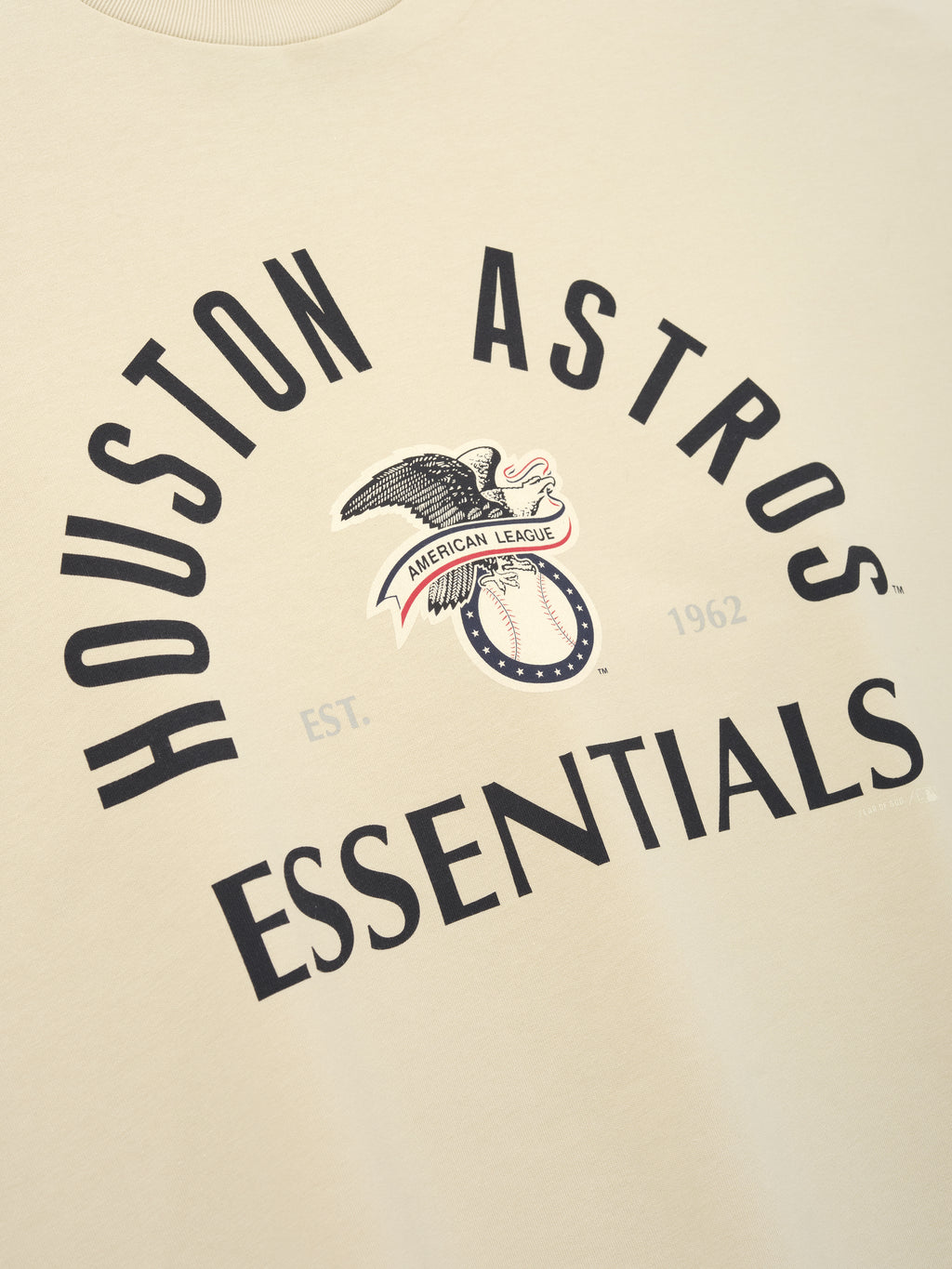 Close-up of a sandstone-colored Astros Al Merch Tee with black curved "Houston Astros Essentials" text and a vintage eagle graphic holding a ribbon reading "American League" on the front.