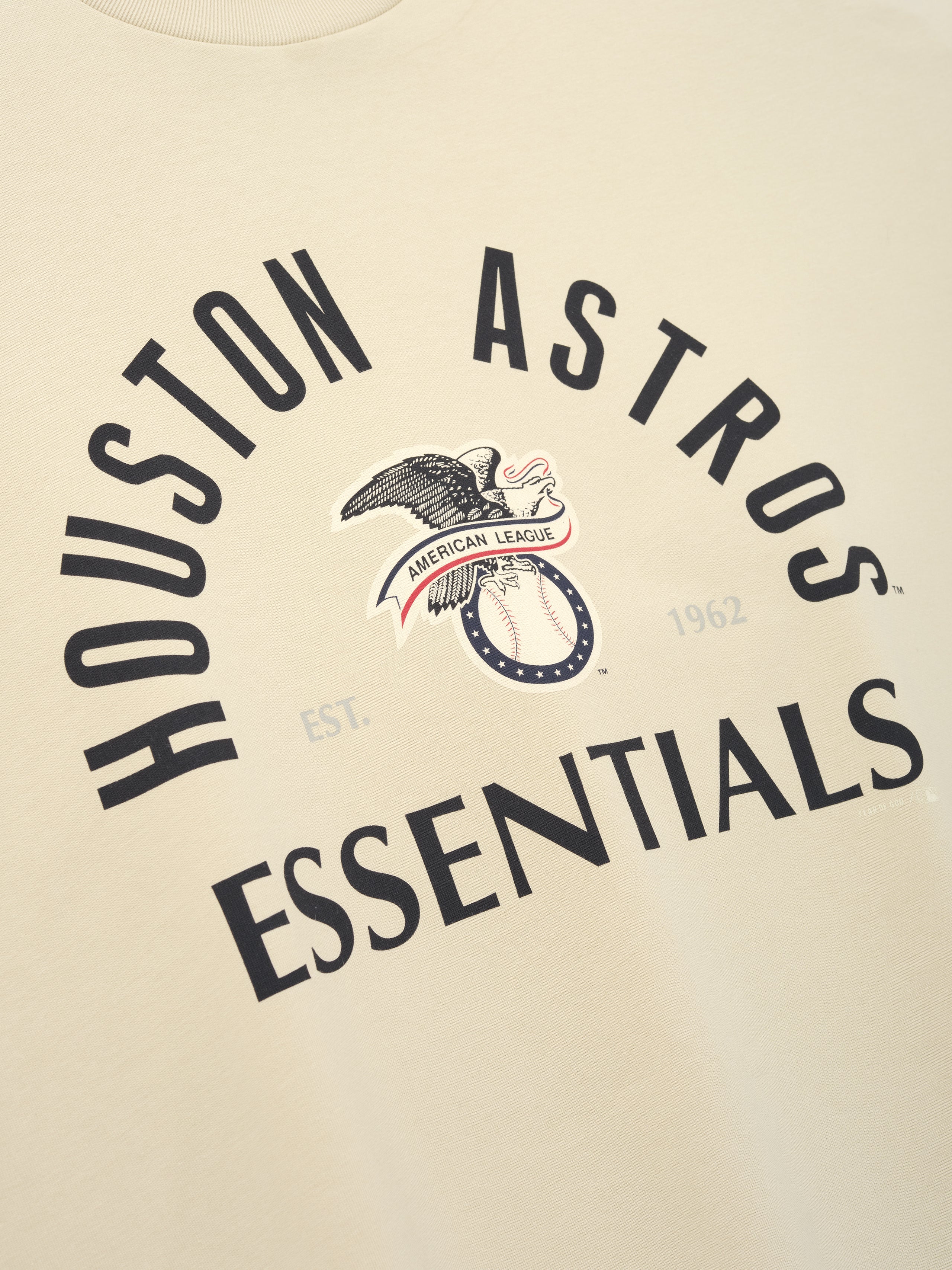 Close-up of a sandstone-colored Astros Al Merch Tee with black curved "Houston Astros Essentials" text and a vintage eagle graphic holding a ribbon reading "American League" on the front.