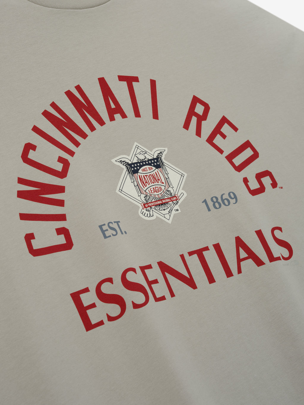 Reds NL Merch Tee in soft beige fabric featuring bold red arched "Cincinnati Reds" text and "Essentials" below, with a vintage-inspired emblem and establishment date in muted blue.
